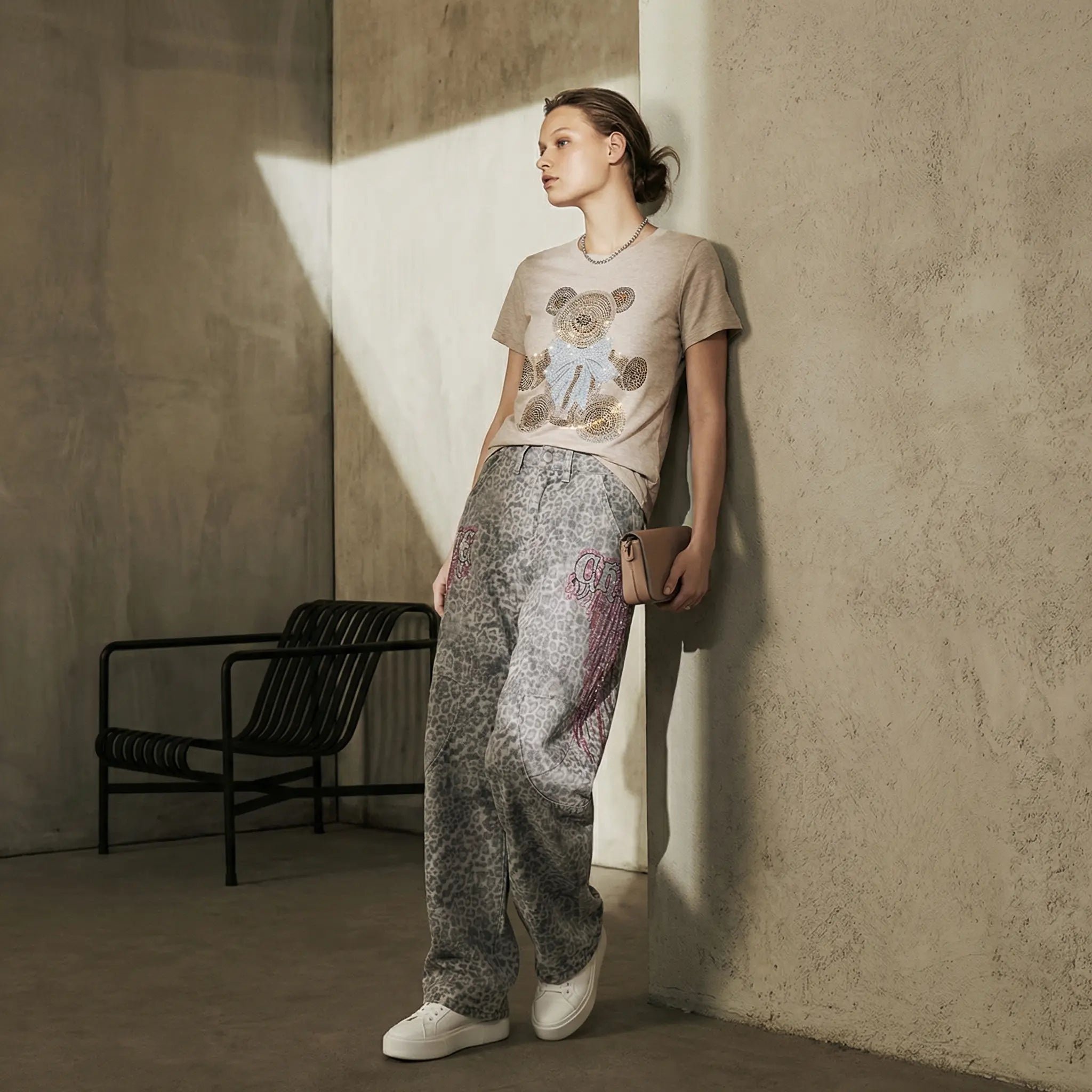 Woman standing against a textured wall wearing a beige t-shirt with a graphic design and patterned pants. TCH Grey Leopard Rhinestone Wings Wide-Leg Jeans
