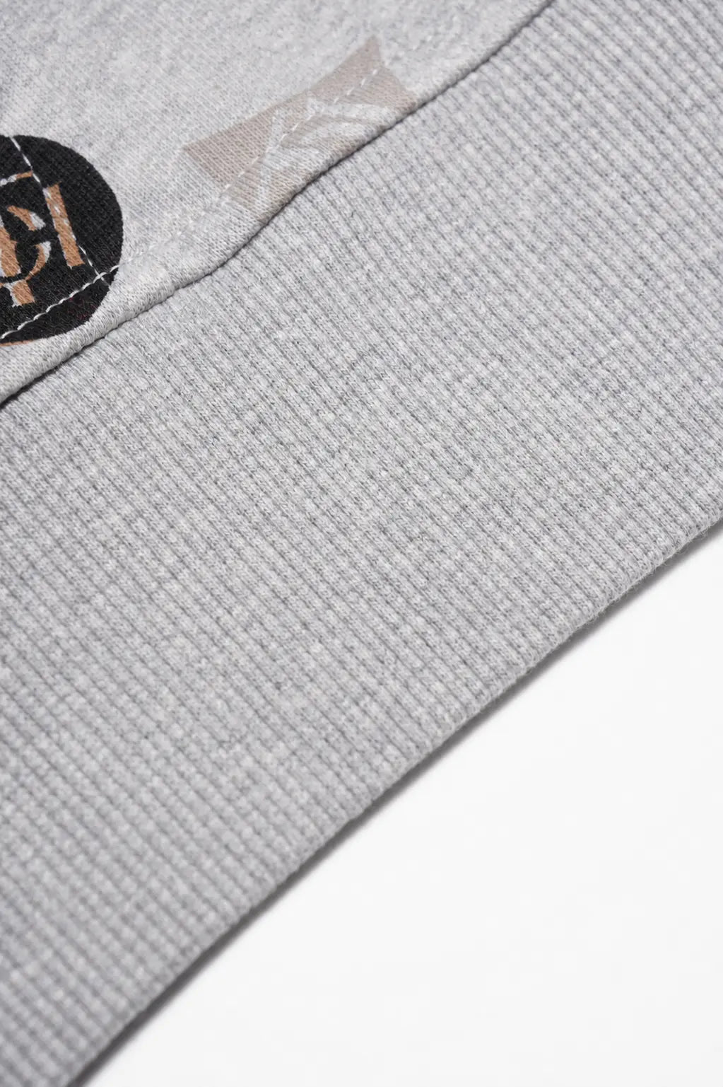 Close-up of a light gray ribbed knit fabric product corner with a small black circular patch and a subtle tonal logo. TCH Grey Logo Print Zip Hoodie