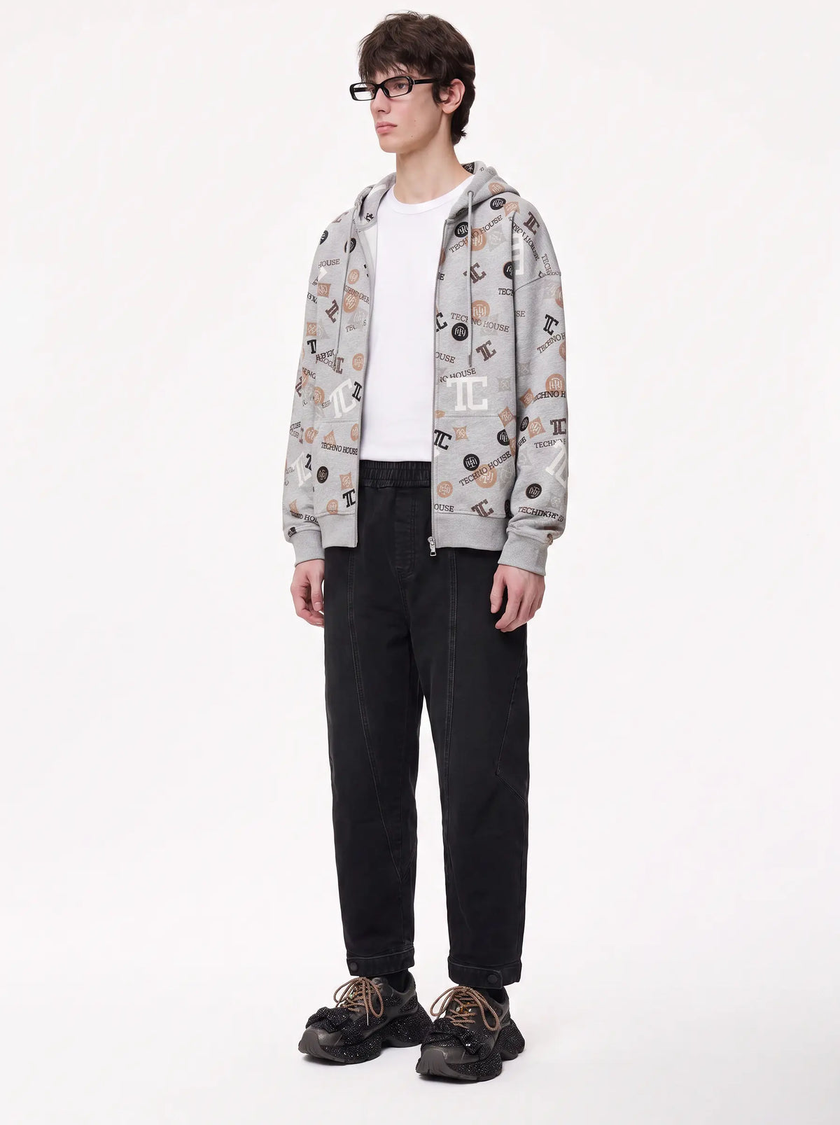 A model wearing a light gray zip-up hoodie with an all-over black and beige graphic print, styled over a white t-shirt, black jeans, and black sneakers, photographed against a plain white studio background. TCH Grey Logo Print Zip Hoodie