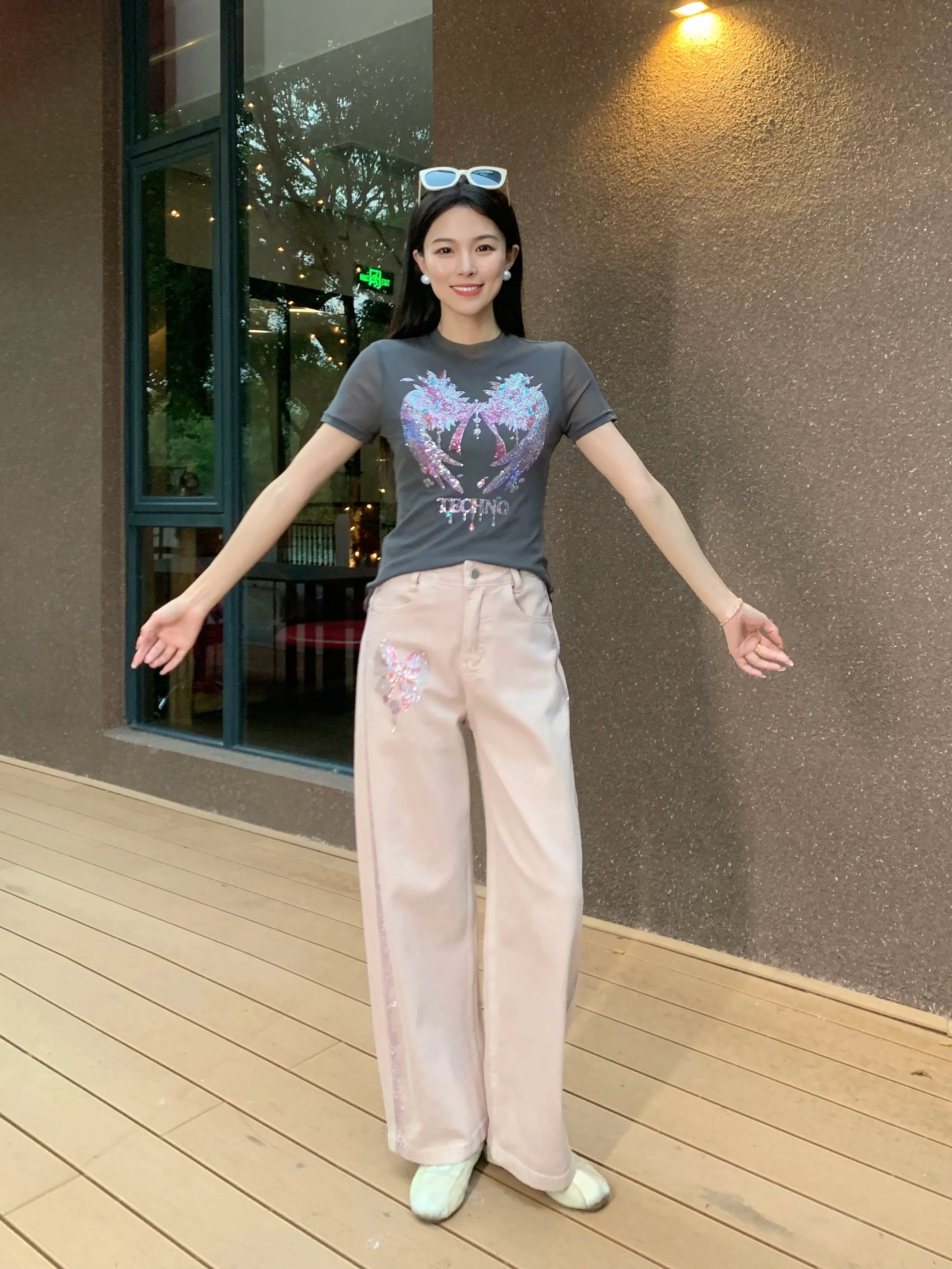 Woman wearing a graphic t-shirt and light-colored pants standing on a wooden deck. TCH Grey Rhinestone Pink Wings Graphic T-Shirt