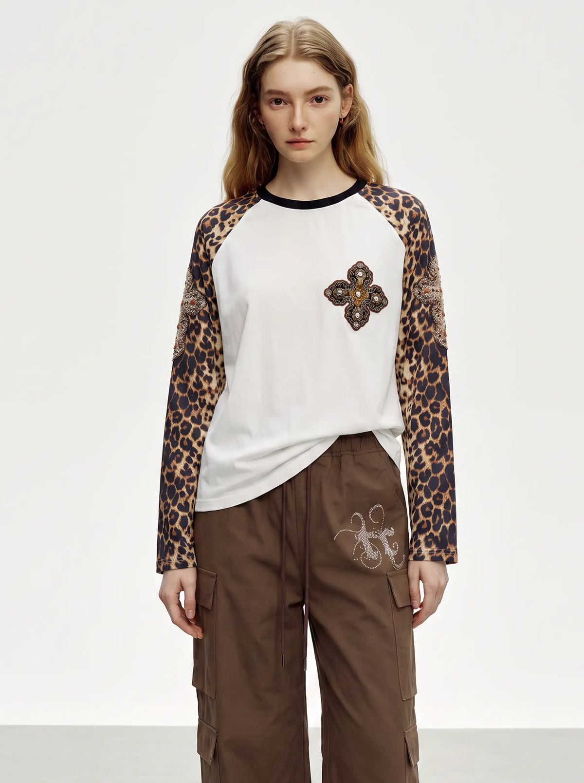 A model wearing a white raglan long-sleeve top with leopard-print sleeves, a black crew neck, and a brown embroidered clover motif on the chest, paired with brown cargo pants against a plain white studio background. TCH Leopard Print Sleeve Embellished Cross Raglan Long Sleeve T-Shirt