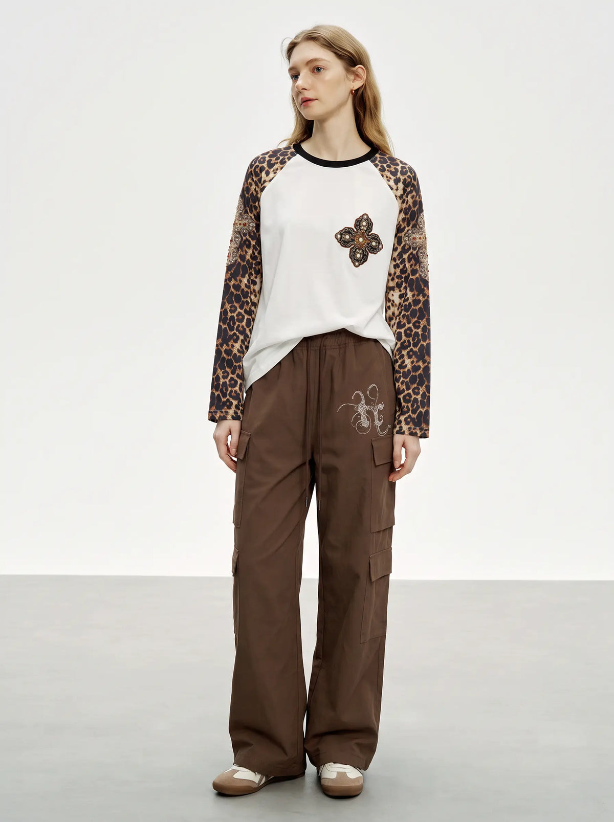 A model stands in a studio wearing a long-sleeve raglan top with leopard-print sleeves, a white body, and a brown embroidered flower motif on the chest. She is also wearing brown wide-leg cargo pants and brown shoes. TCH Leopard Print Sleeve Embellished Cross Raglan Long Sleeve T-Shirt