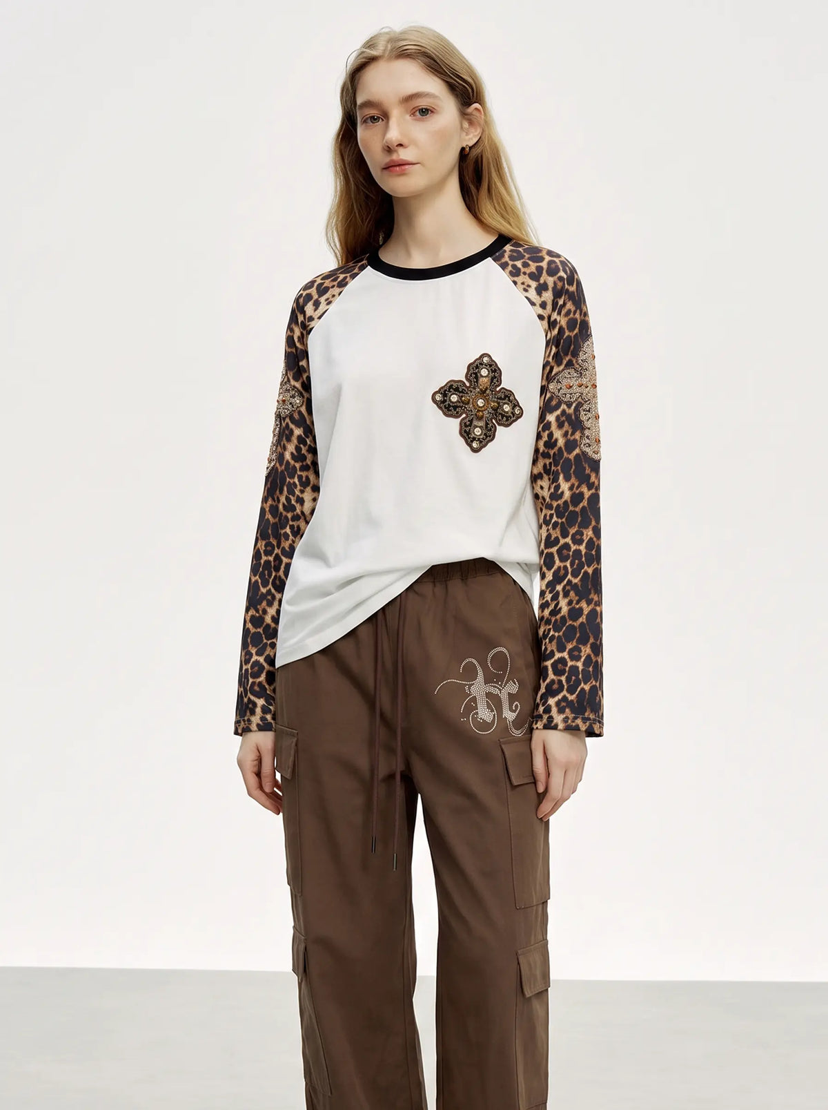 A model stands against a plain light gray studio background wearing a long-sleeve raglan top with leopard-print sleeves, a white body, a black collar, and a floral applique on the chest, paired with brown cargo pants. TCH Leopard Print Sleeve Embellished Cross Raglan Long Sleeve T-Shirt