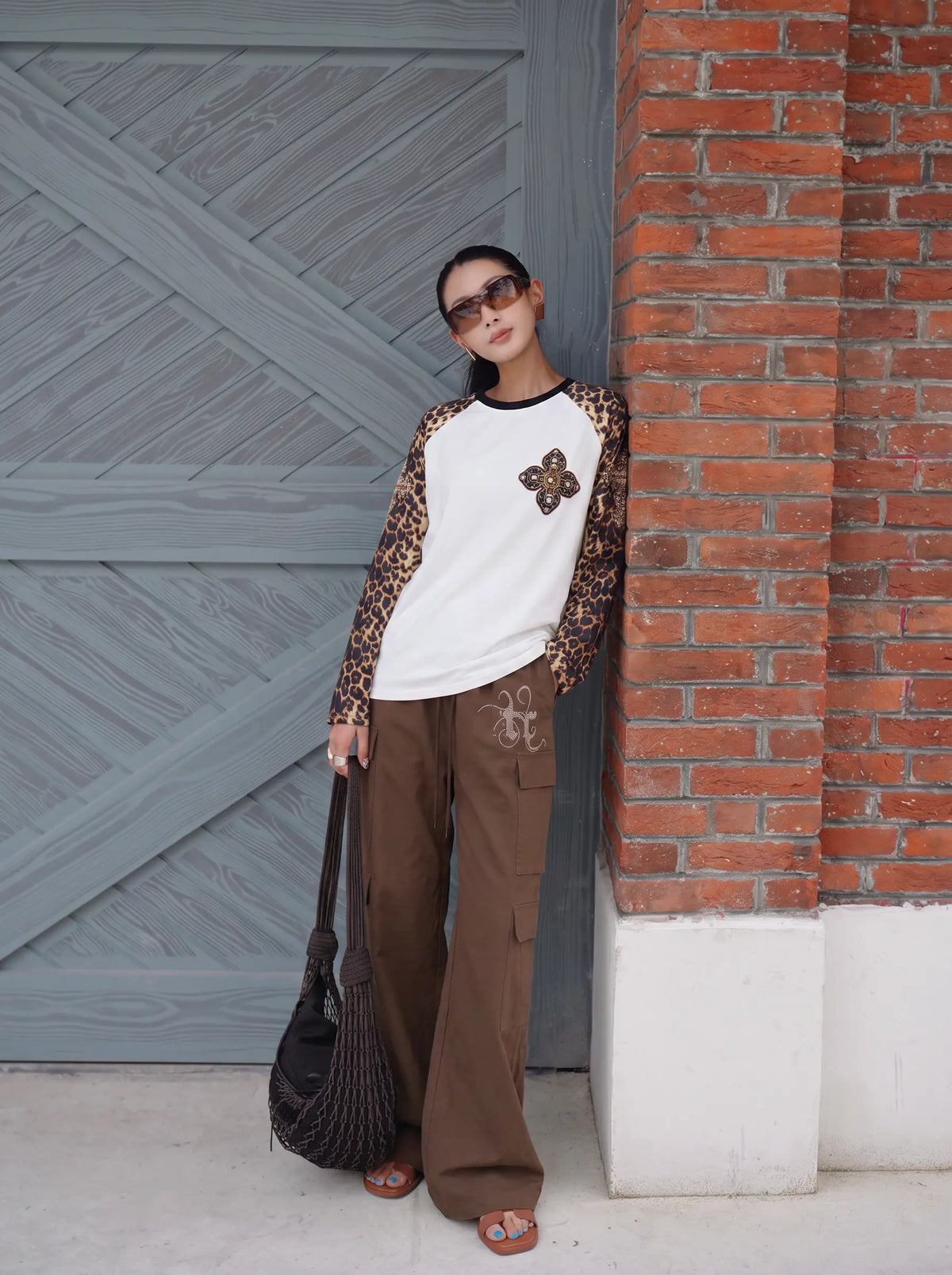 A woman wearing a white and leopard-print raglan long-sleeve shirt, brown wide-leg cargo pants, and sunglasses, holding a black woven handbag and leaning against a brick and gray wooden wall. TCH Leopard Print Sleeve Embellished Cross Raglan Long Sleeve T-Shirt