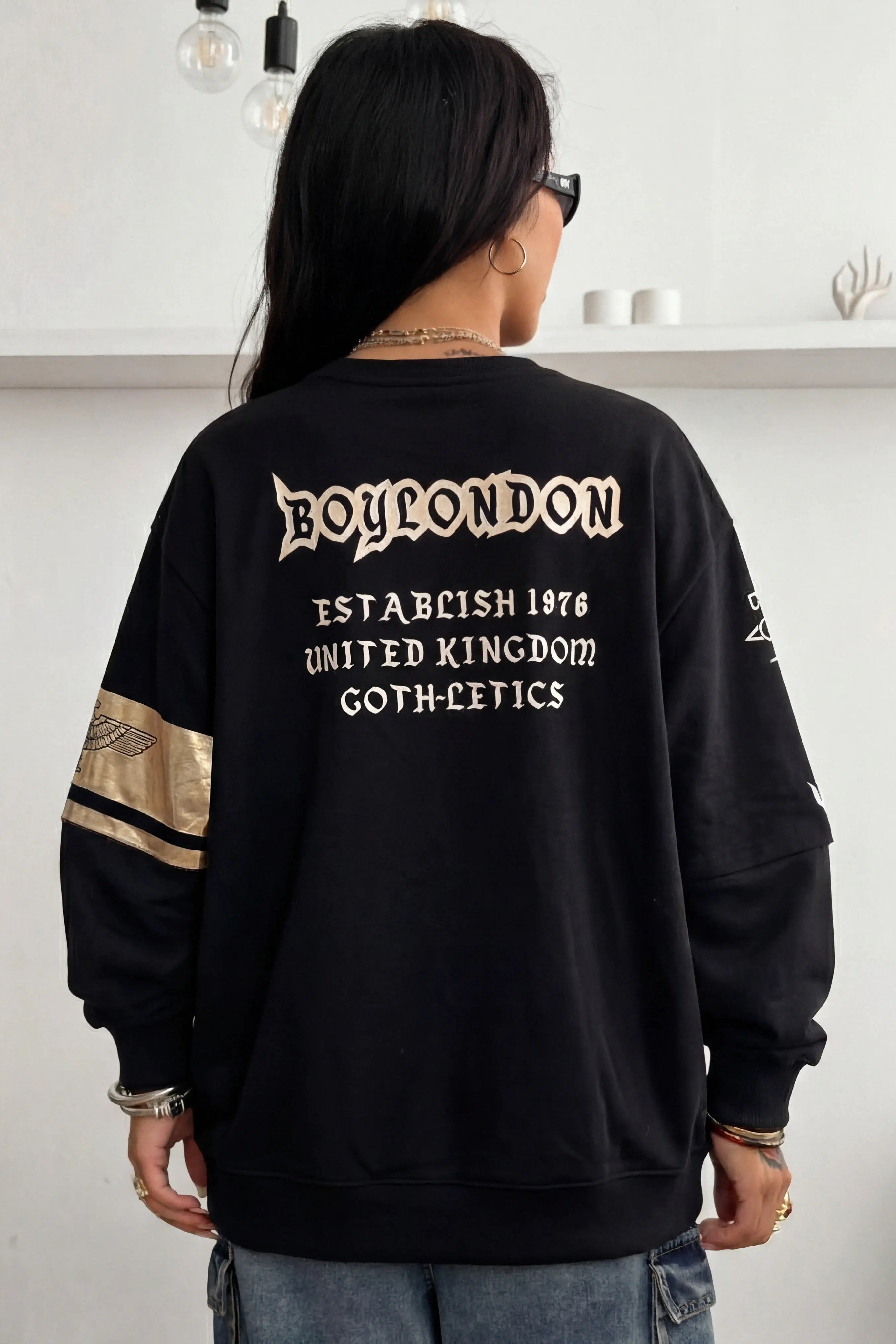 Person wearing a black sweatshirt with text on the back in a minimalistic indoor setting, TCH Lucky Pig Rhinestone Sweatshirt