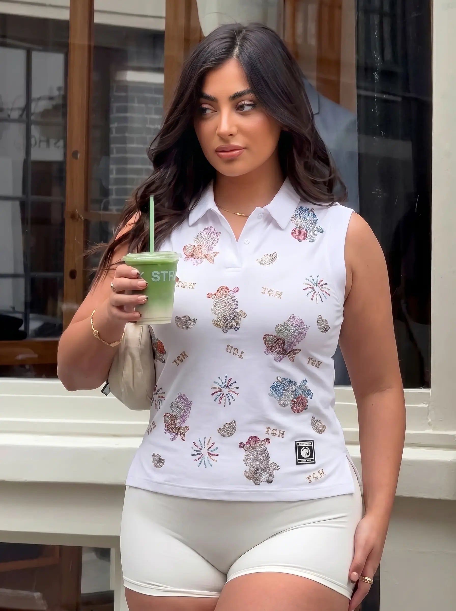 Woman in a white sleeveless floral blouse holding a green drink outside a building. TCH Lucky Pony Rhinestone Sleeveless Polo Shirt