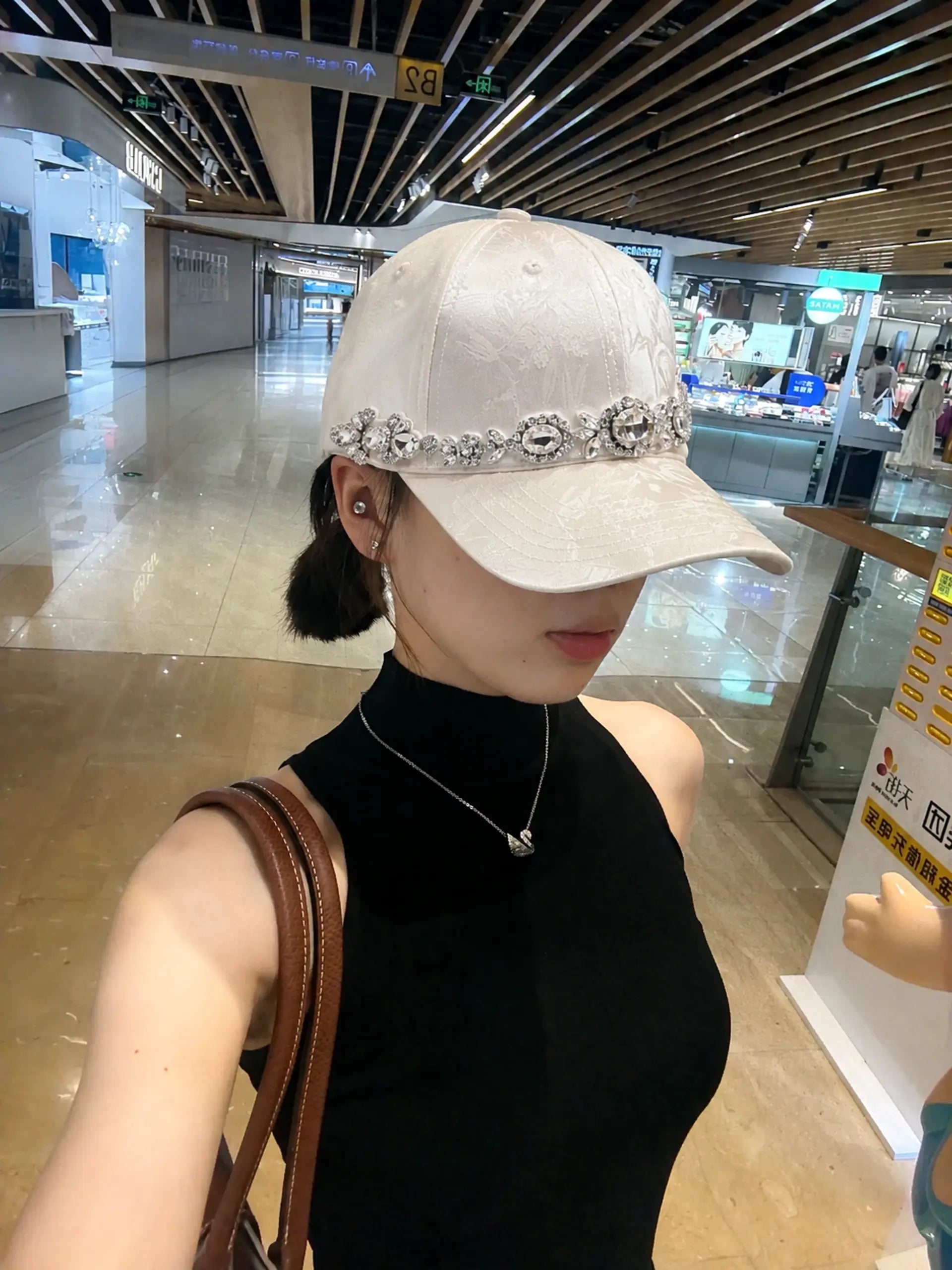 A woman in a black sleeveless top wearing a light beige baseball cap adorned with a silver rhinestone band, photographed in a mall interior. TCH Luxury Crystal Embellished Floral Jacquard Baseball Cap