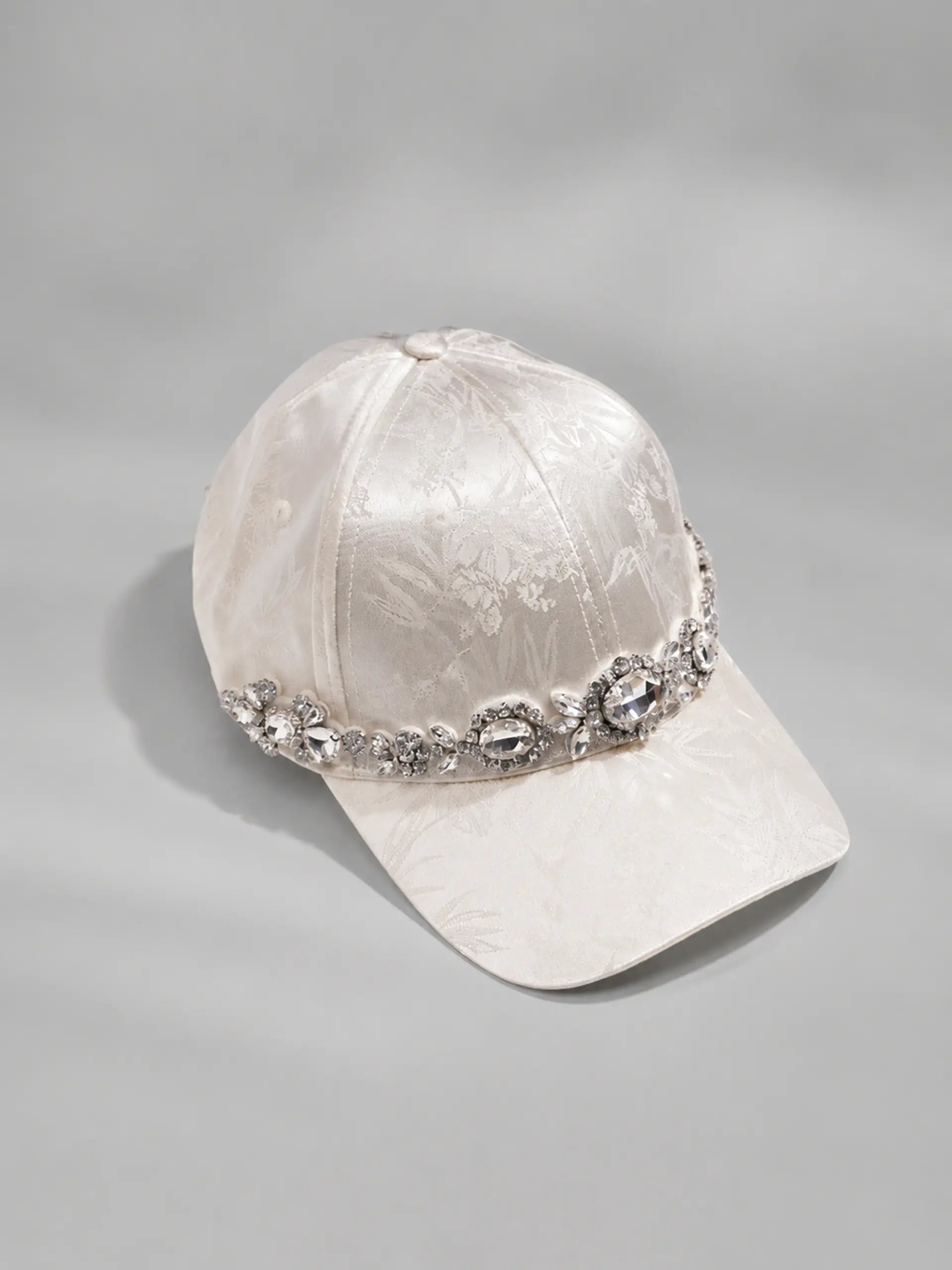 A white jacquard baseball cap adorned with a sparkling silver rhinestone band around the crown, set against a neutral gray background. TCH Luxury Crystal Embellished Floral Jacquard Baseball Cap