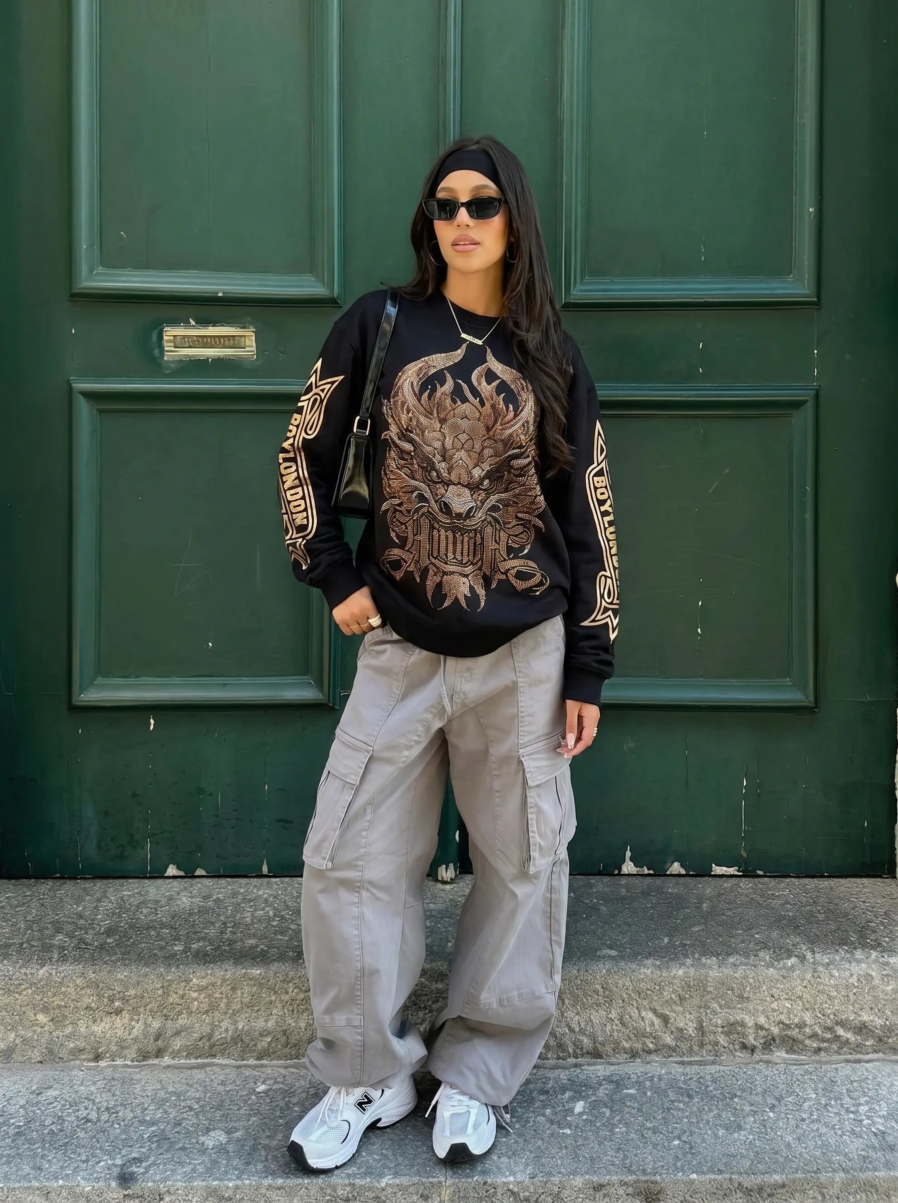 A woman stands in front of a dark green paneled door wearing a black graphic sweatshirt with a large dragon design and gold sleeve lettering, light gray cargo pants, white sneakers, and a black headband. TCH Luxury Gold Dragon Rhinestone Graphic Black Sweatshirt