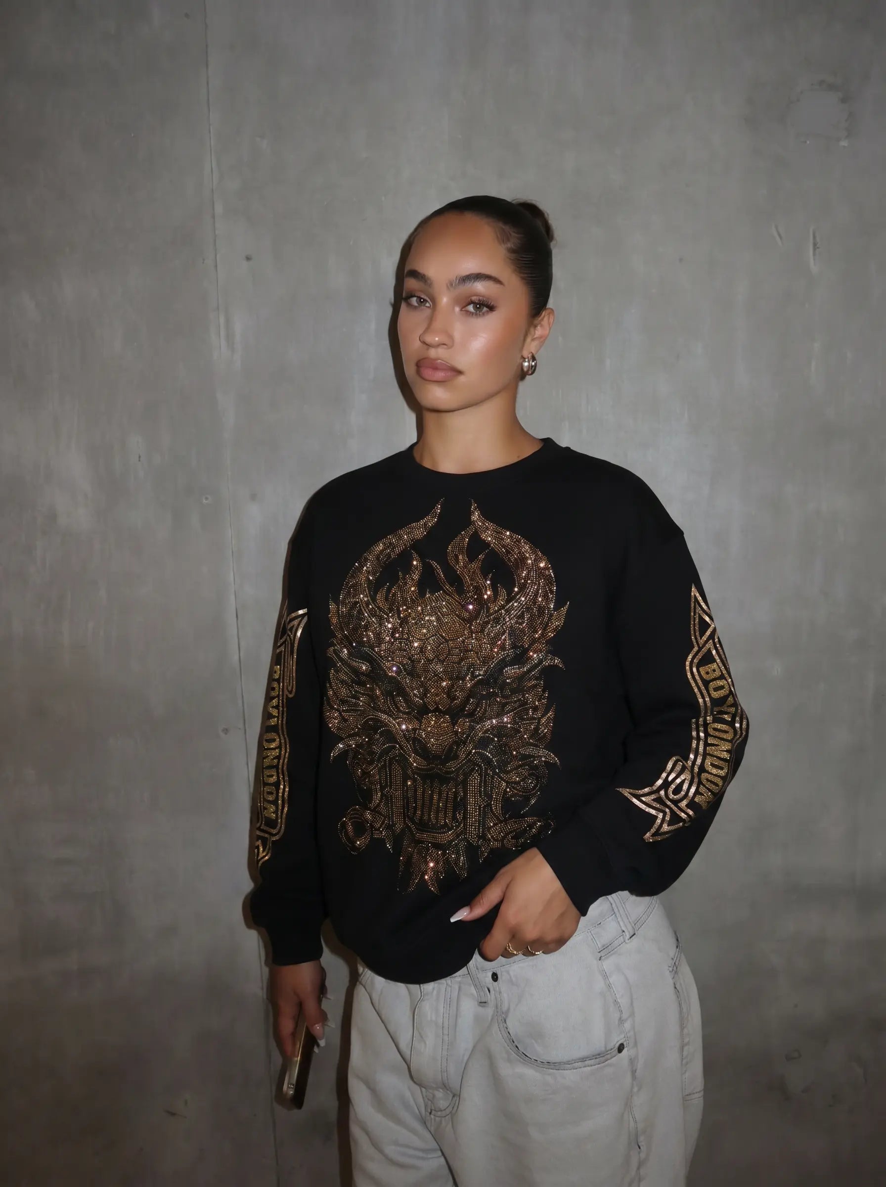 A woman wearing a black crewneck sweatshirt with a large gold and brown metallic graphic on the front and sleeve prints, paired with light gray jeans, standing against a textured gray wall. TCH Luxury Gold Dragon Rhinestone Graphic Black Sweatshirt