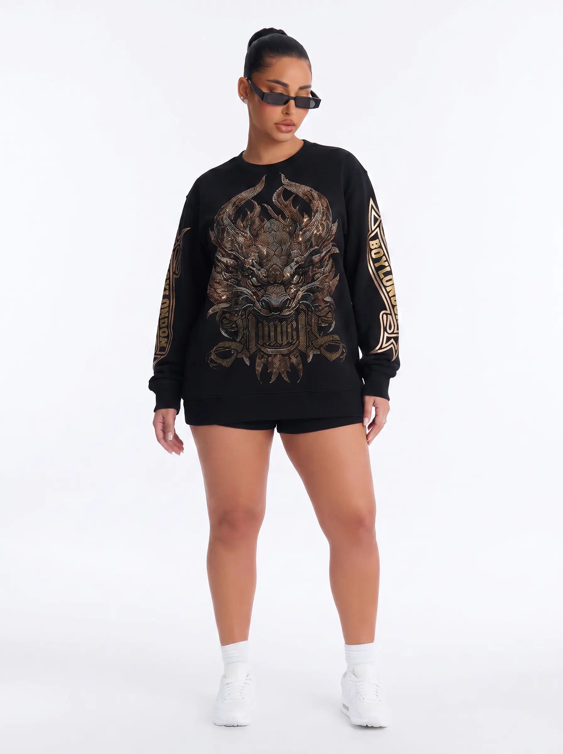 A model wearing a black crewneck sweatshirt with a large metallic gold dragon graphic on the front and gold lettering down the sleeves, paired with black shorts, white socks, and black sunglasses against a white studio background. TCH Luxury Gold Dragon Rhinestone Graphic Black Sweatshirt