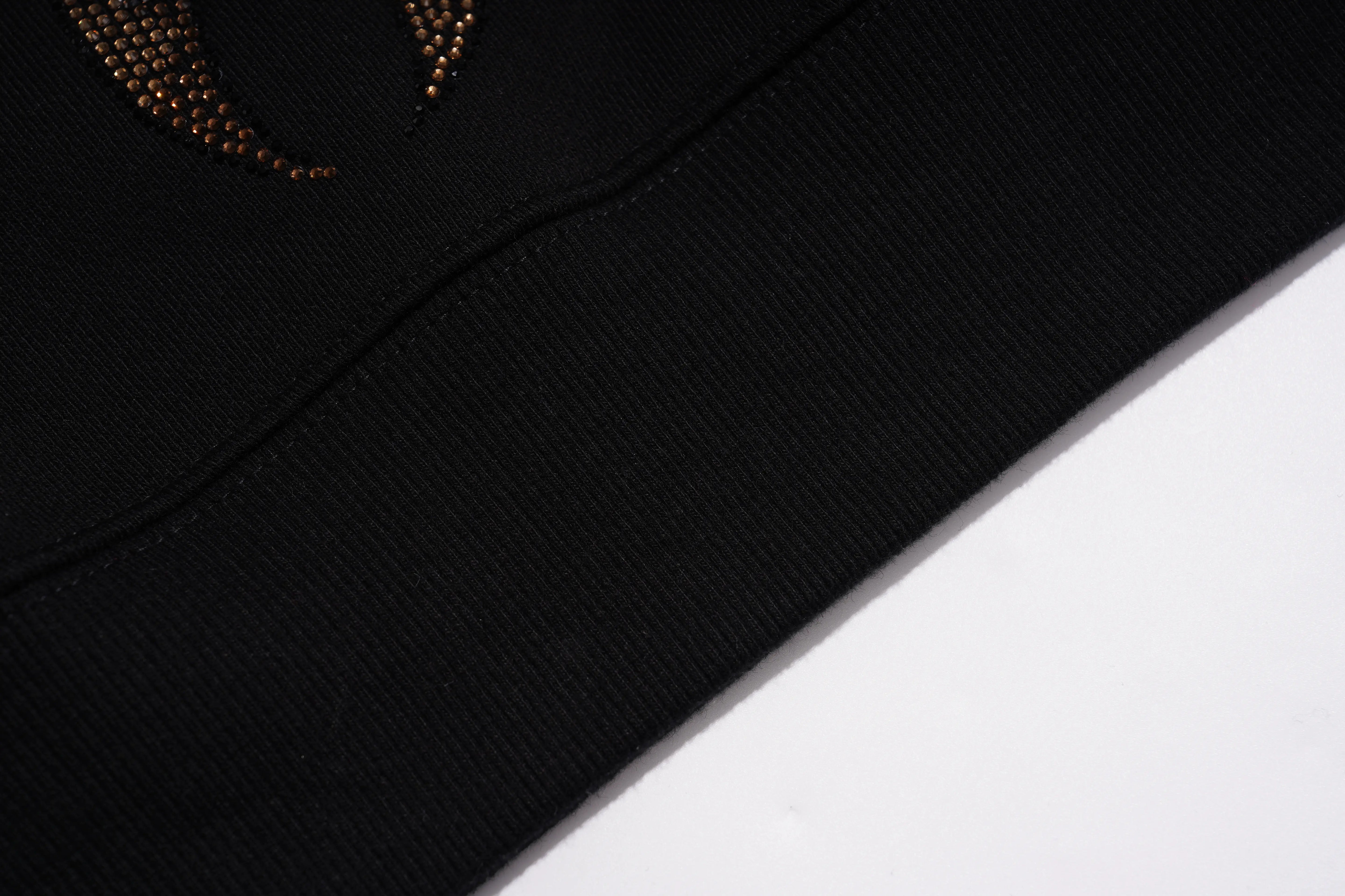 Close-up detail of a black ribbed knit garment, showing a curved seam and a small section of a metallic gold beaded embellishment on the upper left, set against a plain white background. TCH Luxury Gold Dragon Rhinestone Graphic Black Sweatshirt