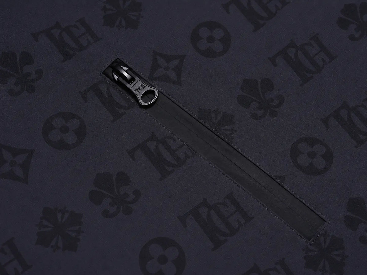 A close-up of a black zipper with a dark metal pull, placed on a dark navy fabric background patterned with a repeating Louis Vuitton monogram. TCH Luxury Monogram Hooded Windbreaker Jacket