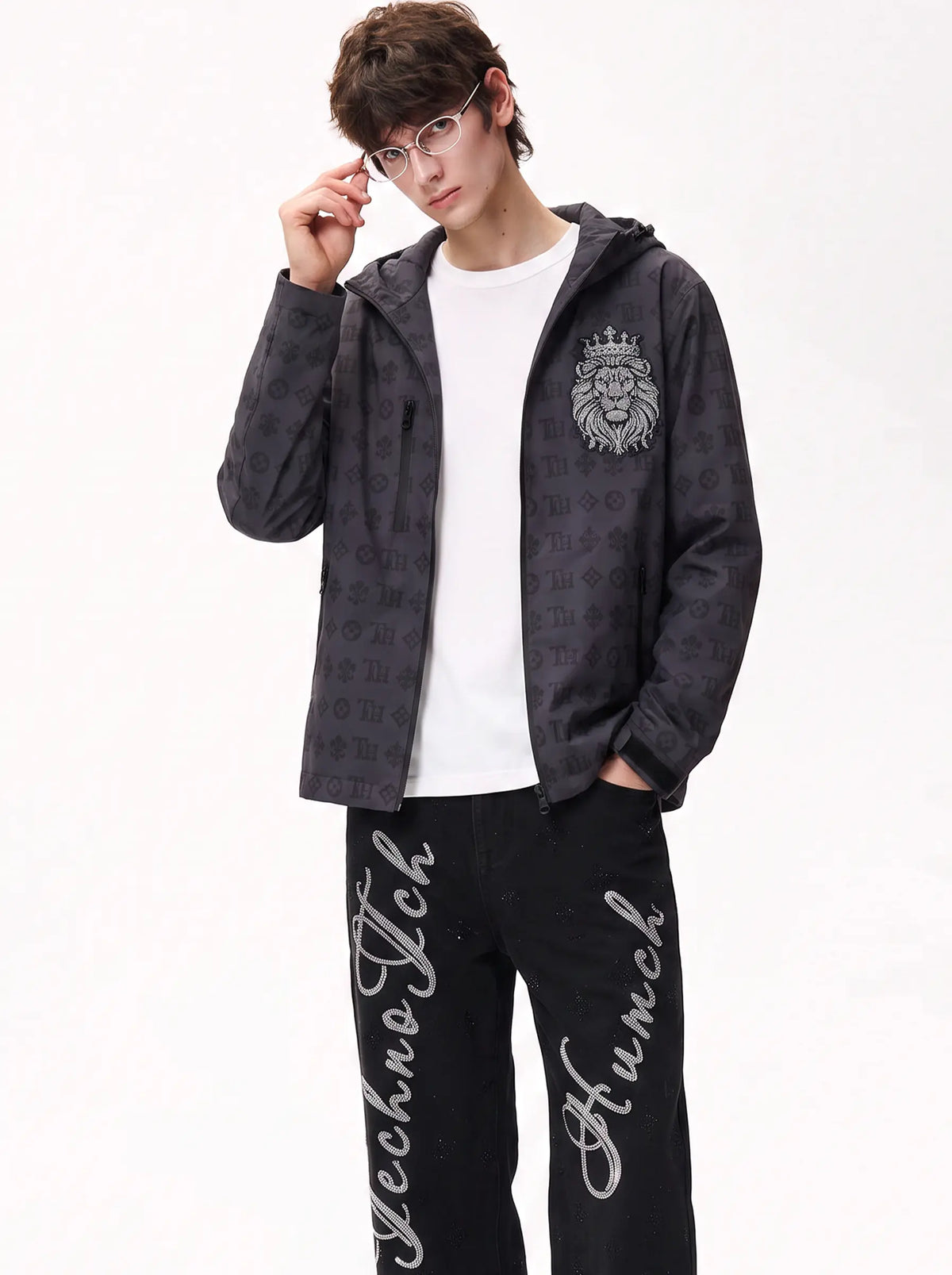 A man wearing a dark gray patterned zip-up hoodie with a lion crest on the chest, a white t-shirt, and black sweatpants with white script text, posed against a plain white background. TCH Luxury Monogram Hooded Windbreaker Jacket