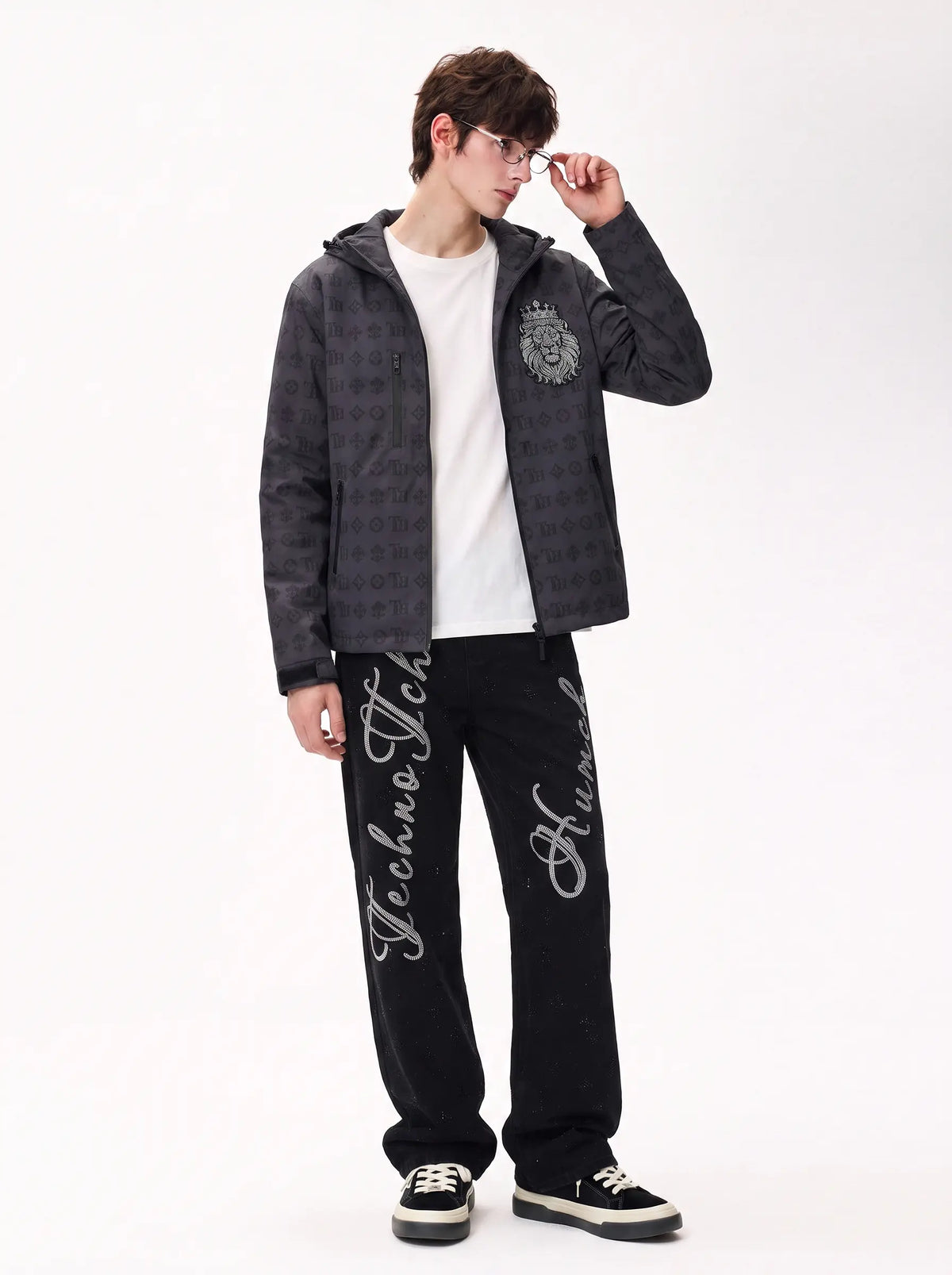 A model wearing a dark gray patterned hooded jacket, a white t-shirt, black pants with white script embroidery, and black sneakers, standing against a plain white studio background. TCH Luxury Monogram Hooded Windbreaker Jacket