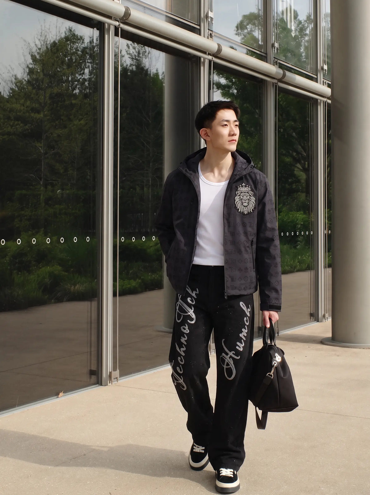 A man walking outdoors in front of a modern glass building, wearing a dark gray zip-up hoodie with a crest logo, a white t-shirt, black sweatpants with white script lettering, and carrying a black duffel bag. TCH Luxury Monogram Hooded Windbreaker Jacket