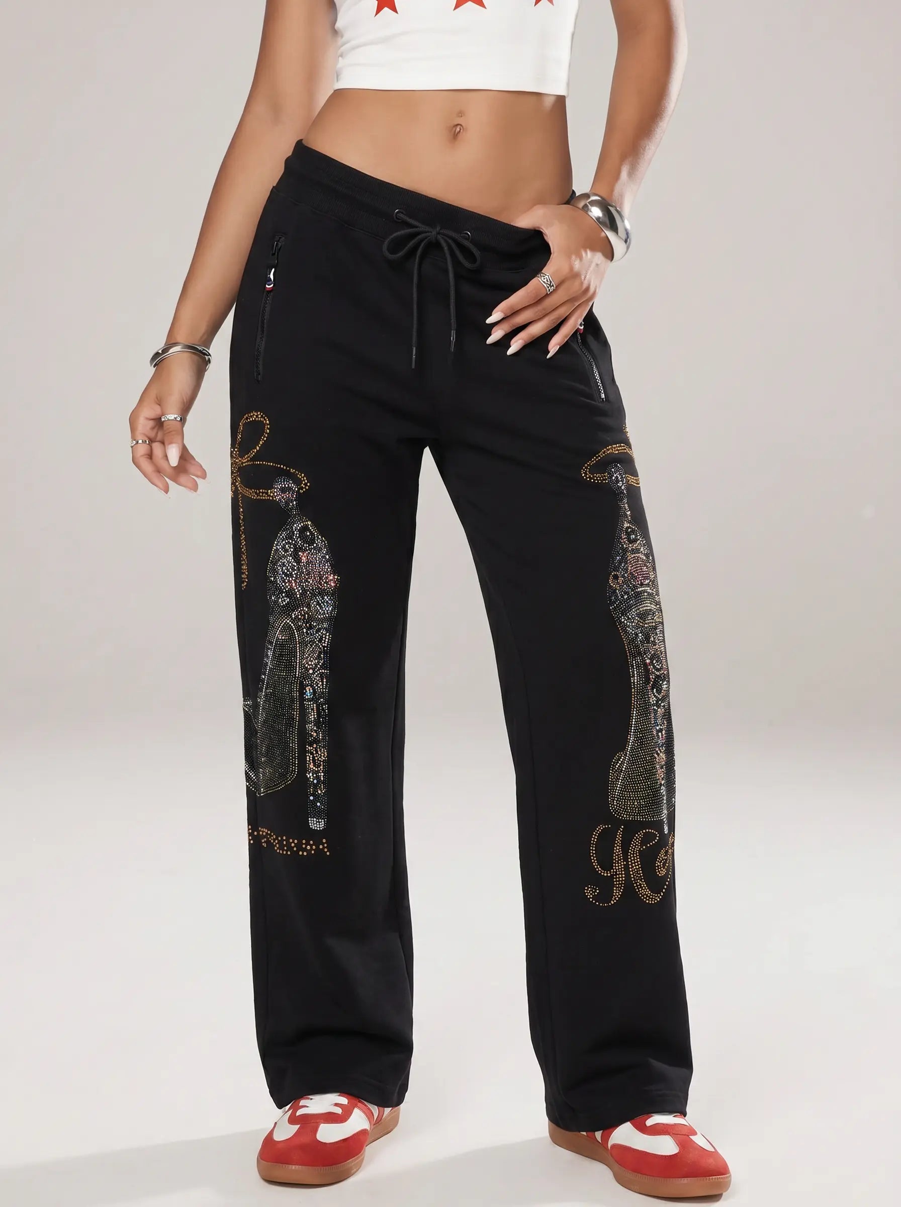 A model wearing black wide-leg sweatpants with gothic-style rhinestone and metallic embroidery, paired with a white crop top and red sneakers, against a light gray studio background. TCH Luxury Rhinestone High Heel Graphic Black Sweatpants