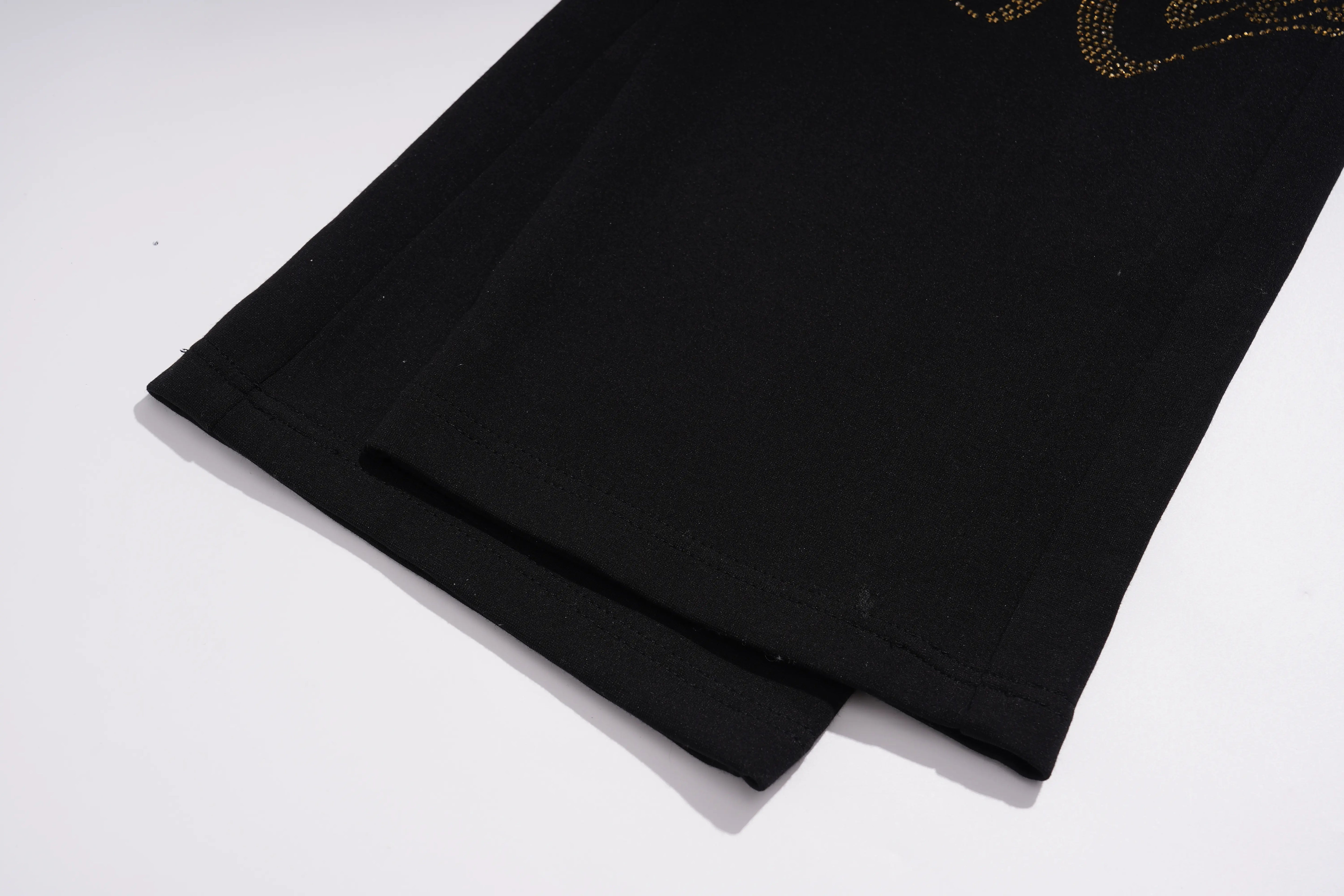 Close-up of a black garment laid flat on a white background, showing the hem and a small portion of a gold-toned graphic near the upper right edge. TCH Luxury Rhinestone High Heel Graphic Black Sweatpants