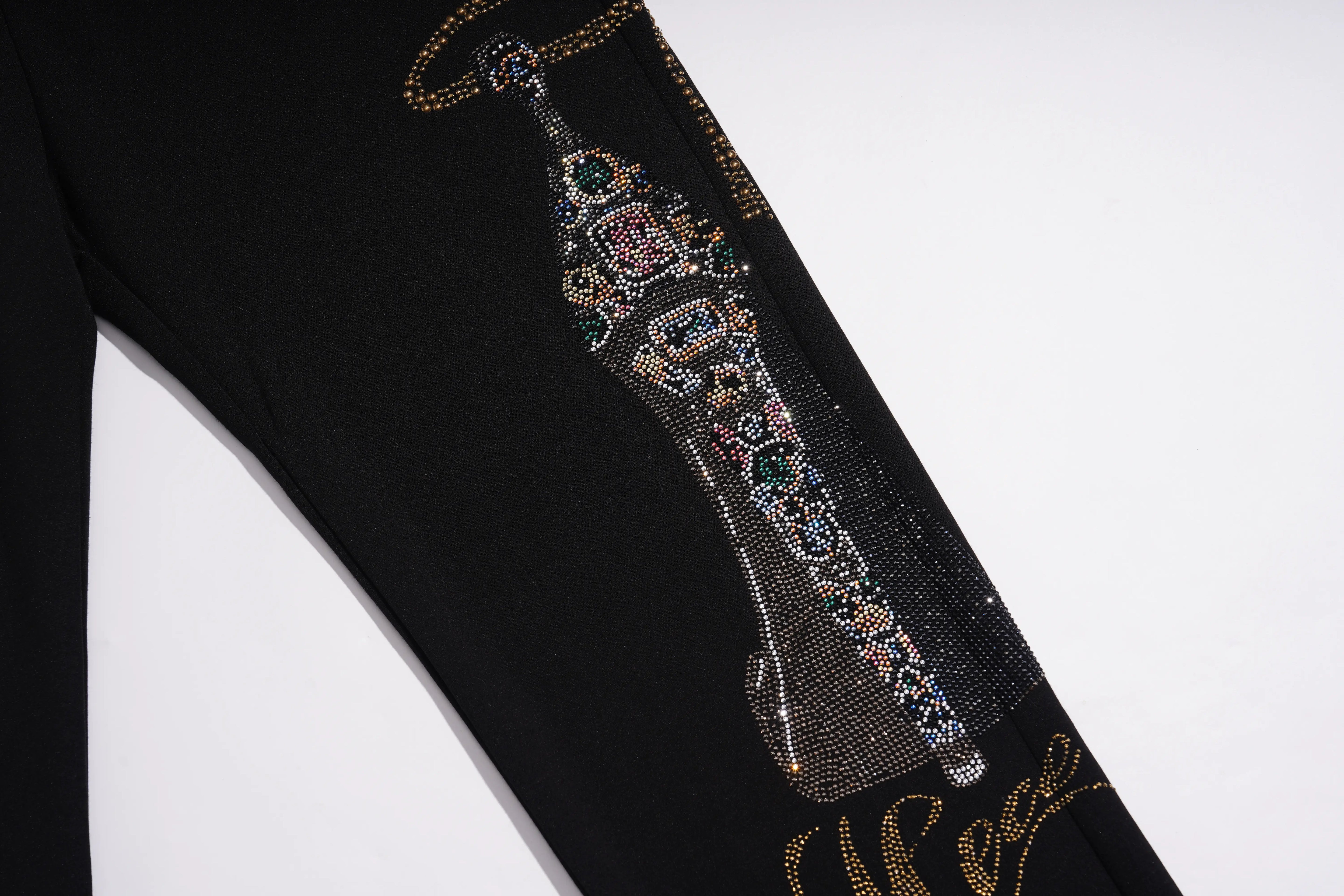 Close-up of a black garment featuring a large, ornate rhinestone embellishment in the center and gold script lettering near the bottom, set against a plain white background. TCH Luxury Rhinestone High Heel Graphic Black Sweatpants