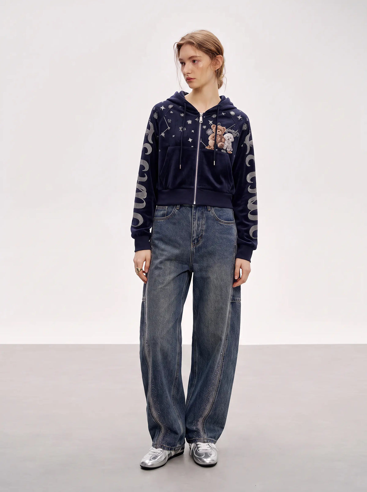 A model stands in a studio wearing a navy blue zip-up hoodie with star patterns, a cartoon graphic on the chest, and gray lettering down the sleeves, paired with wide-leg blue jeans and silver sneakers. TCH Navy Teddy Bear Rhinestone Zip Hoodie