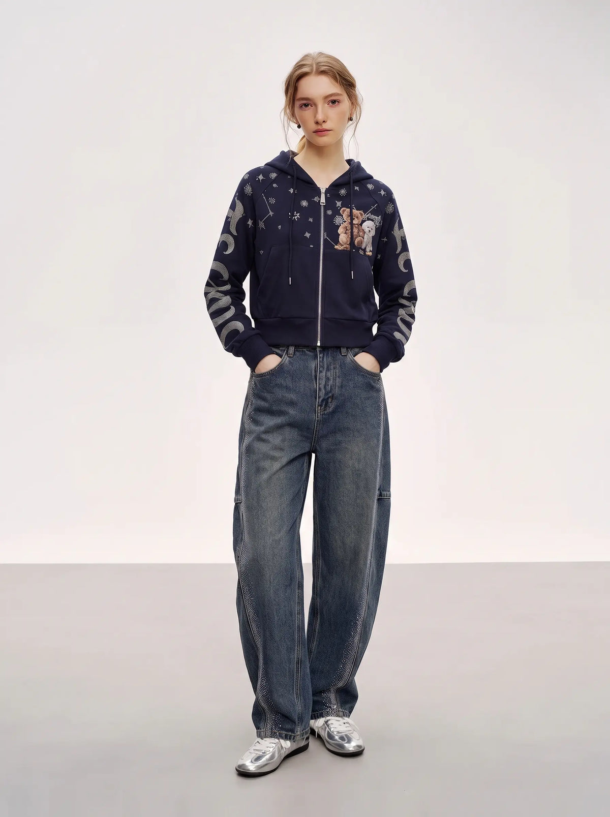 A model stands against a light gray studio background wearing a navy blue zip-up hoodie with celestial and floral embroidery, paired with wide-leg blue jeans and silver sneakers. TCH Navy Teddy Bear Rhinestone Zip Hoodie
