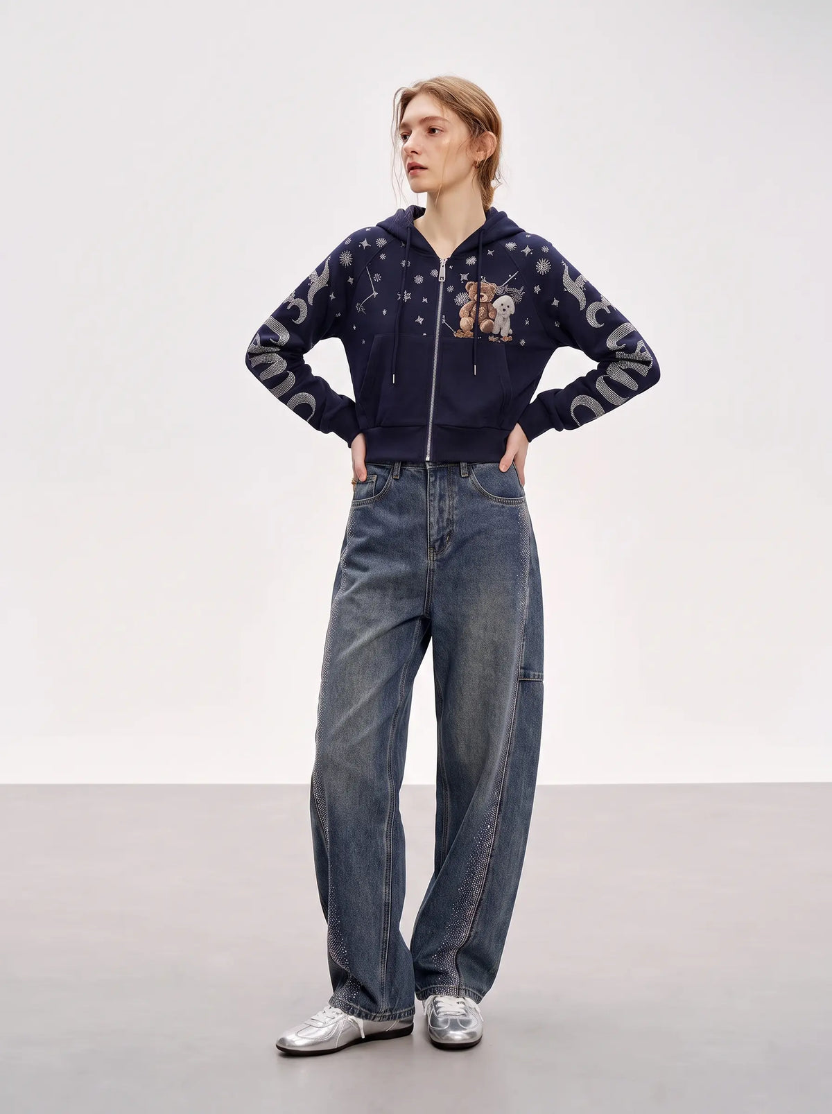 A model stands in a studio wearing a navy blue zip-up hoodie with celestial and cartoon graphics, paired with wide-leg blue jeans and silver sneakers. TCH Navy Teddy Bear Rhinestone Zip Hoodie
