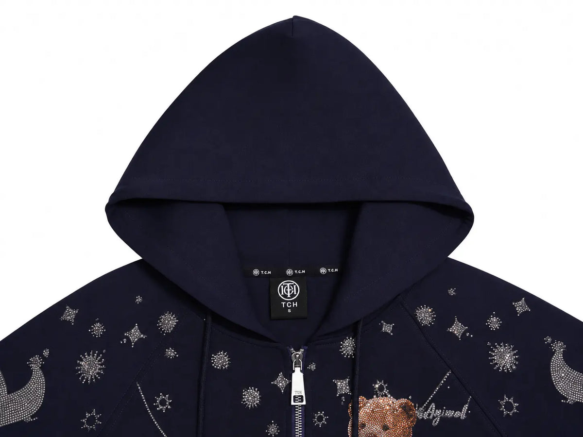 Close-up of a navy blue zip-up hoodie with a hood, featuring silver rhinestone moon and star prints on the chest, a silver zipper, and a teddy bear graphic on the lower front. TCH Navy Teddy Bear Rhinestone Zip Hoodie