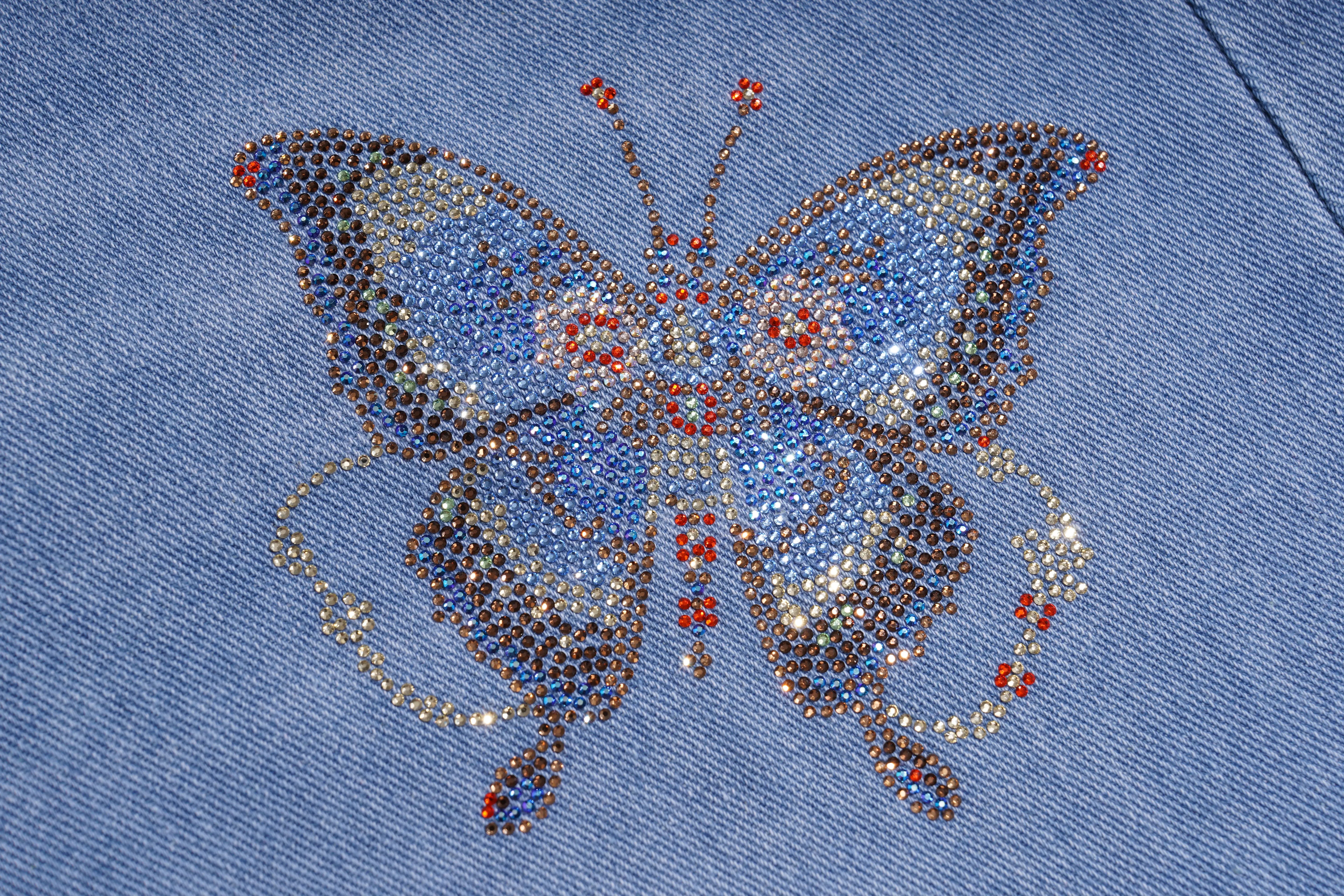 A close-up of a blue denim garment featuring a large, detailed butterfly embellishment made of rhinestones and crystals in shades of blue, silver, and red. TCH Phoenix & Butterfly Rhinestone Wide Leg Jeans