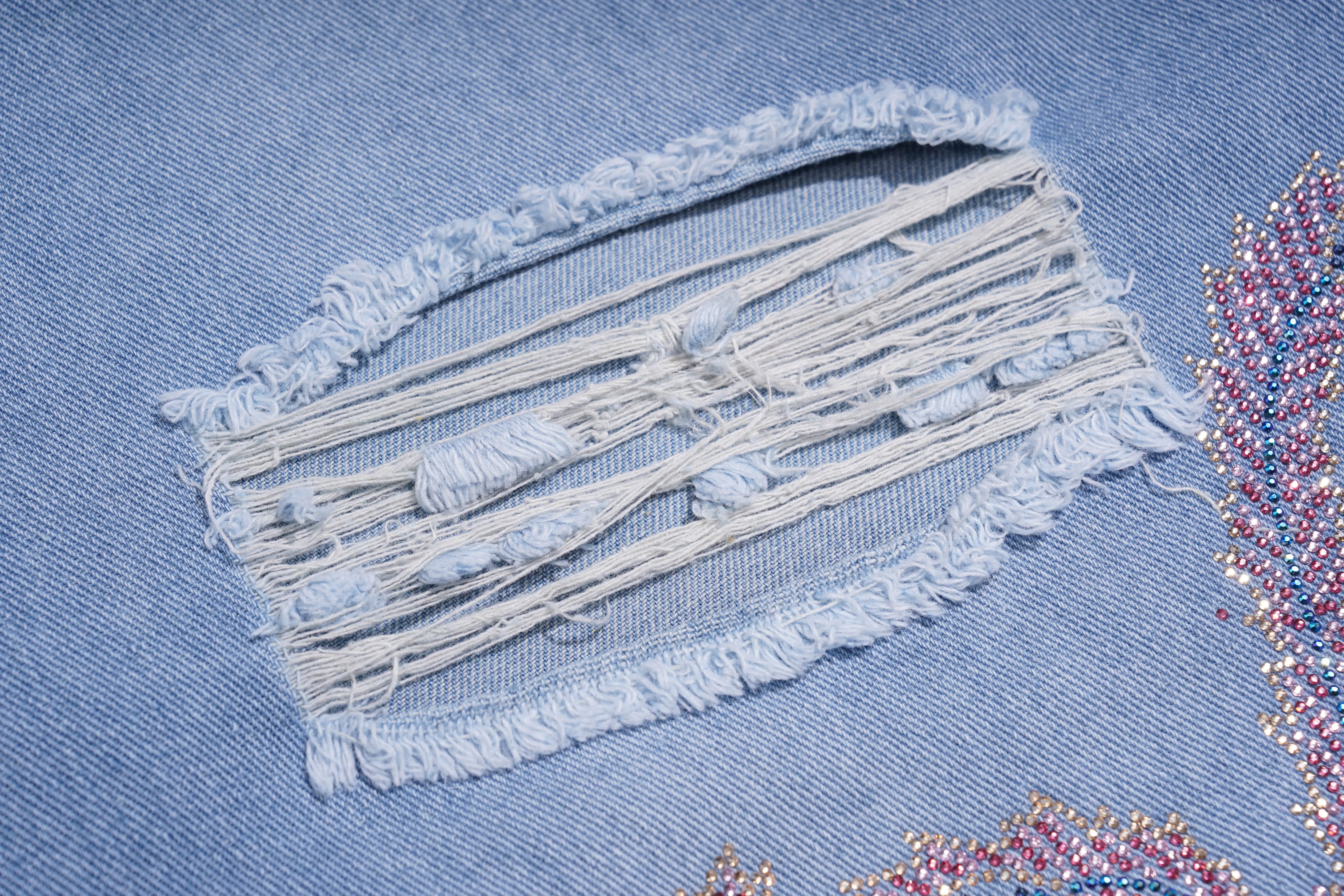 A close-up of a light blue denim garment showing a distressed patch with frayed edges and exposed thread, alongside colorful sequined embroidery. TCH Phoenix & Butterfly Rhinestone Wide Leg Jeans