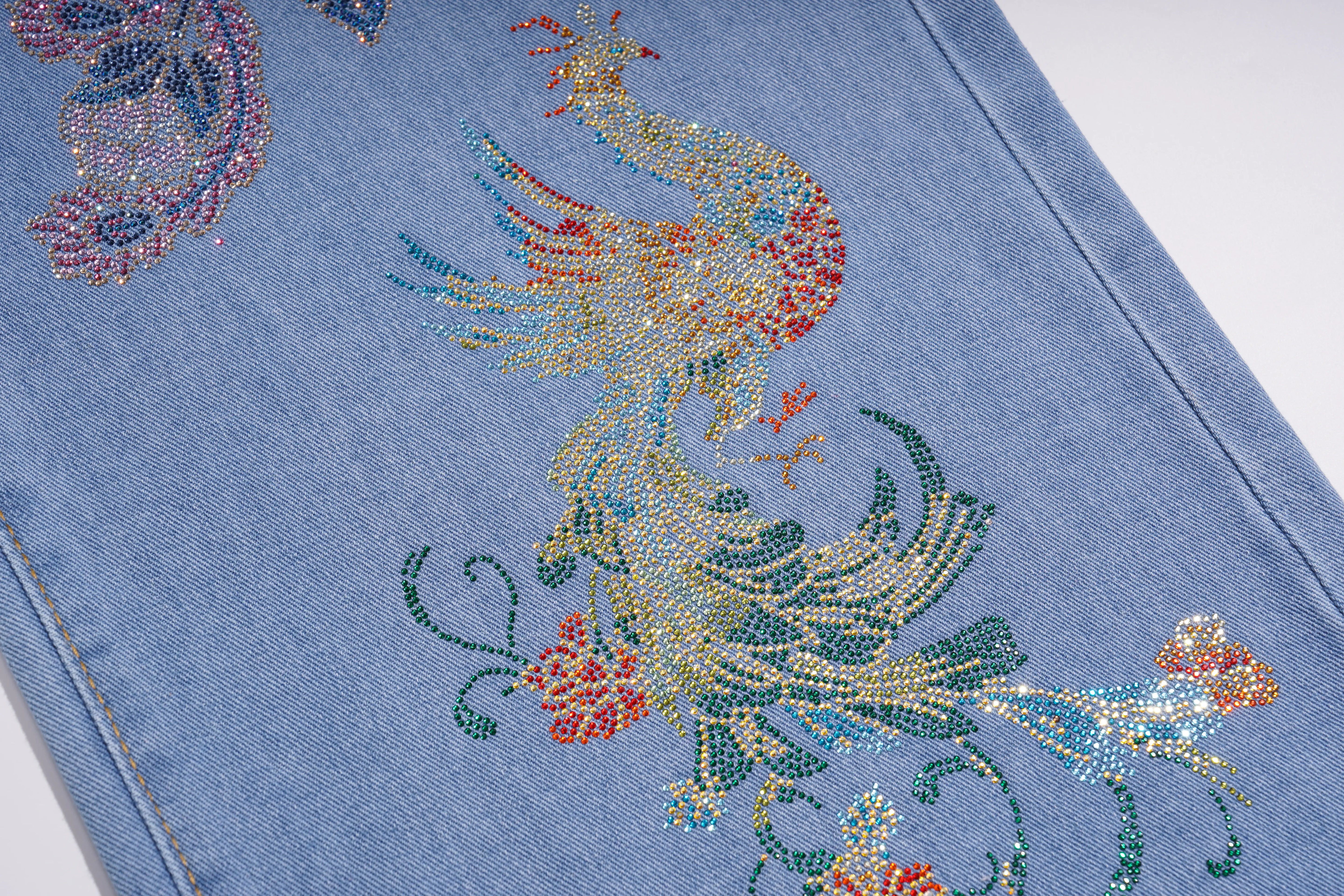 Close-up of a light blue denim garment featuring a large, colorful rhinestone-embellished peacock and floral design on the leg. TCH Phoenix & Butterfly Rhinestone Wide Leg Jeans