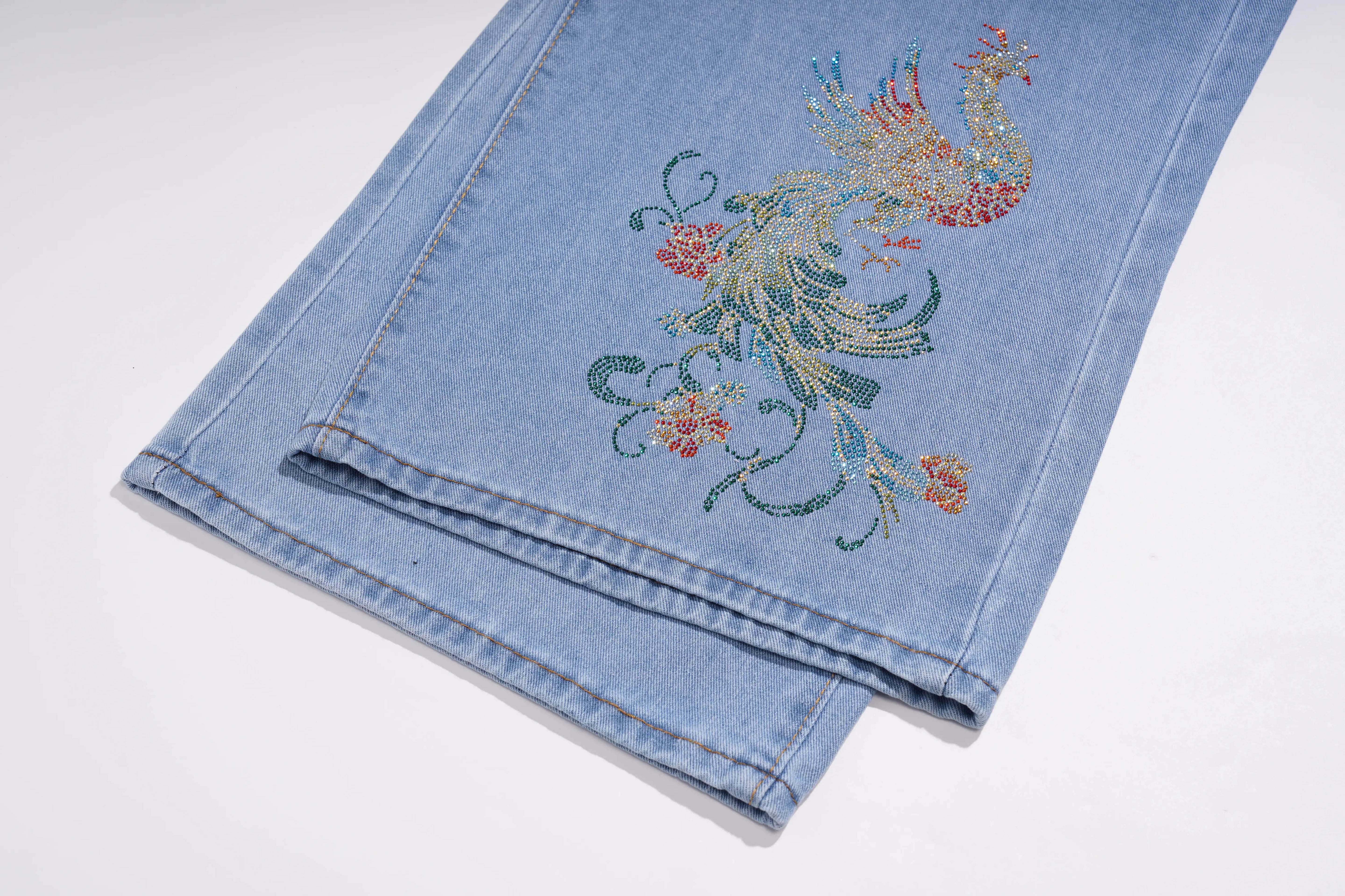 Light blue denim jeans folded on a white background, featuring a colorful embroidered phoenix design with floral details on the leg. TCH Phoenix & Butterfly Rhinestone Wide Leg Jeans