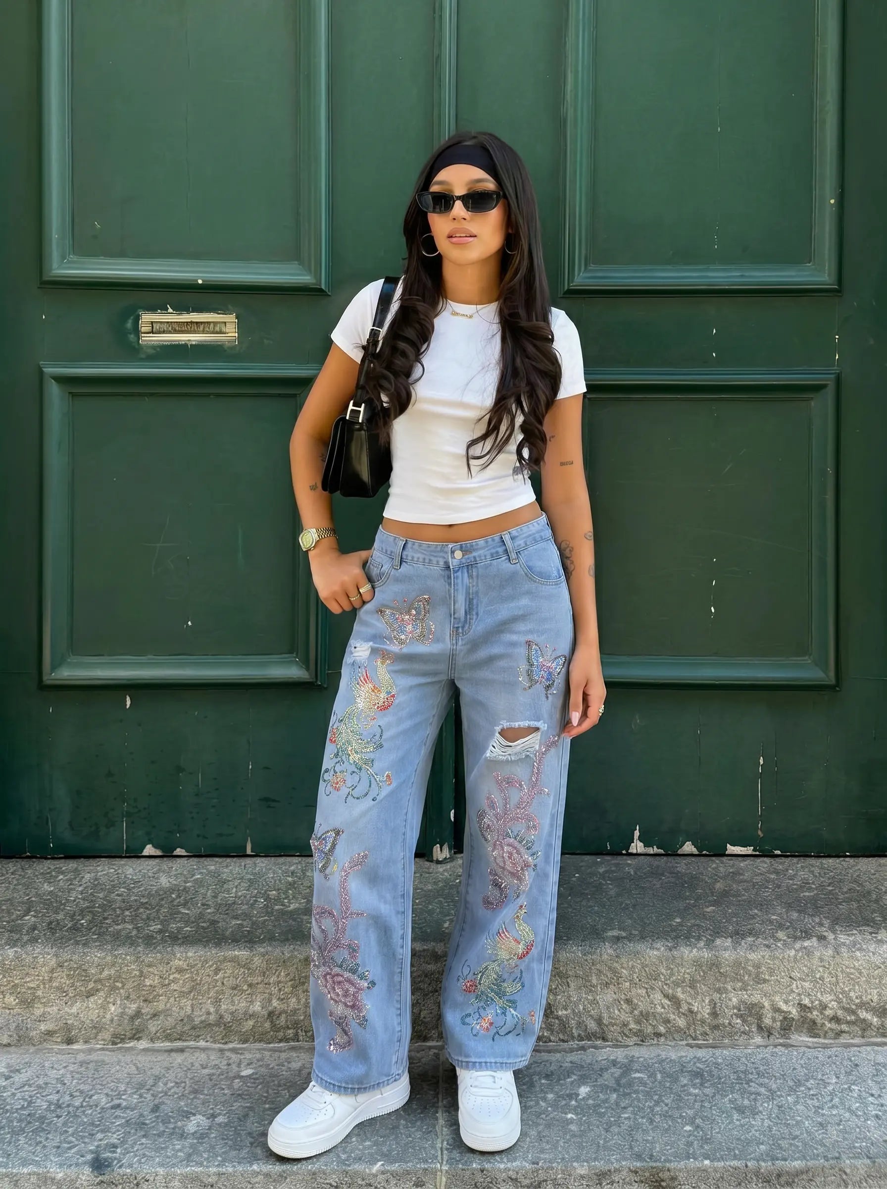 A woman stands outdoors in front of a dark green paneled door wearing light-wash distressed jeans with colorful floral embroidery on the legs, a white cropped T-shirt, white sneakers, sunglasses, and a black shoulder bag. TCH Phoenix & Butterfly Rhinestone Wide Leg Jeans
