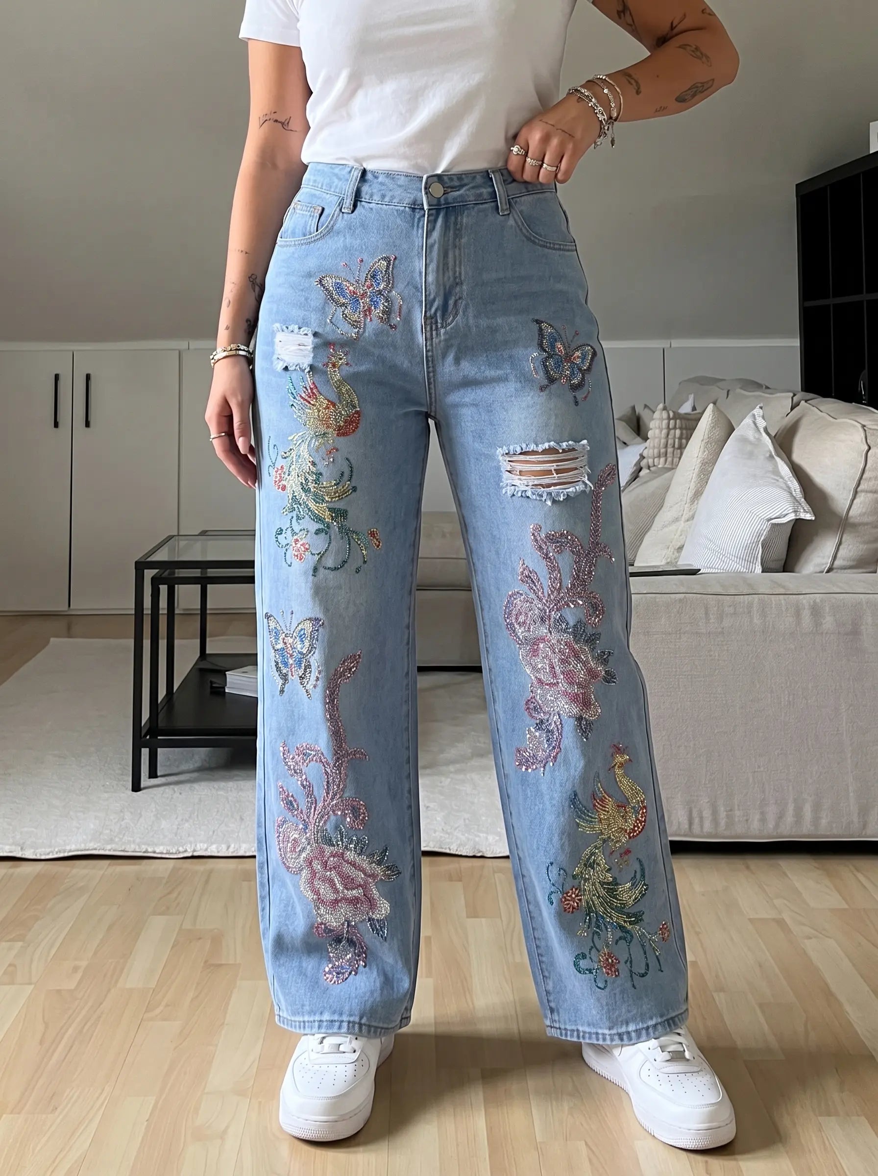 A person wearing light blue wide-leg jeans with colorful embroidered floral and butterfly designs, paired with a white t-shirt and white sneakers in a living room setting. TCH Phoenix & Butterfly Rhinestone Wide Leg Jeans