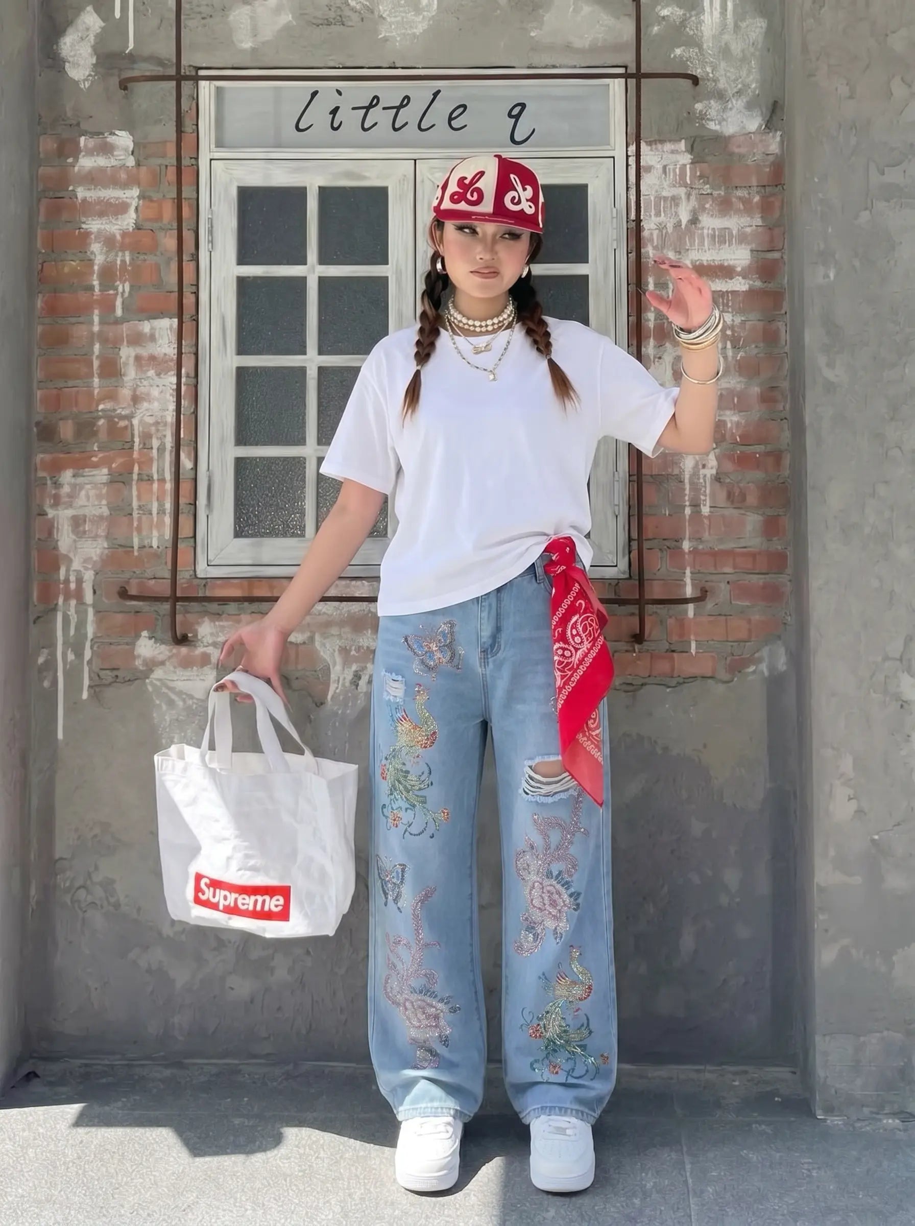 A person stands outdoors in front of a brick and concrete wall, wearing a white T-shirt, light blue distressed jeans with colorful floral embroidery, a red bandana tied around the waist, and a red cap. They are holding a white tote bag with a red Supreme logo. TCH Phoenix & Butterfly Rhinestone Wide Leg Jeans