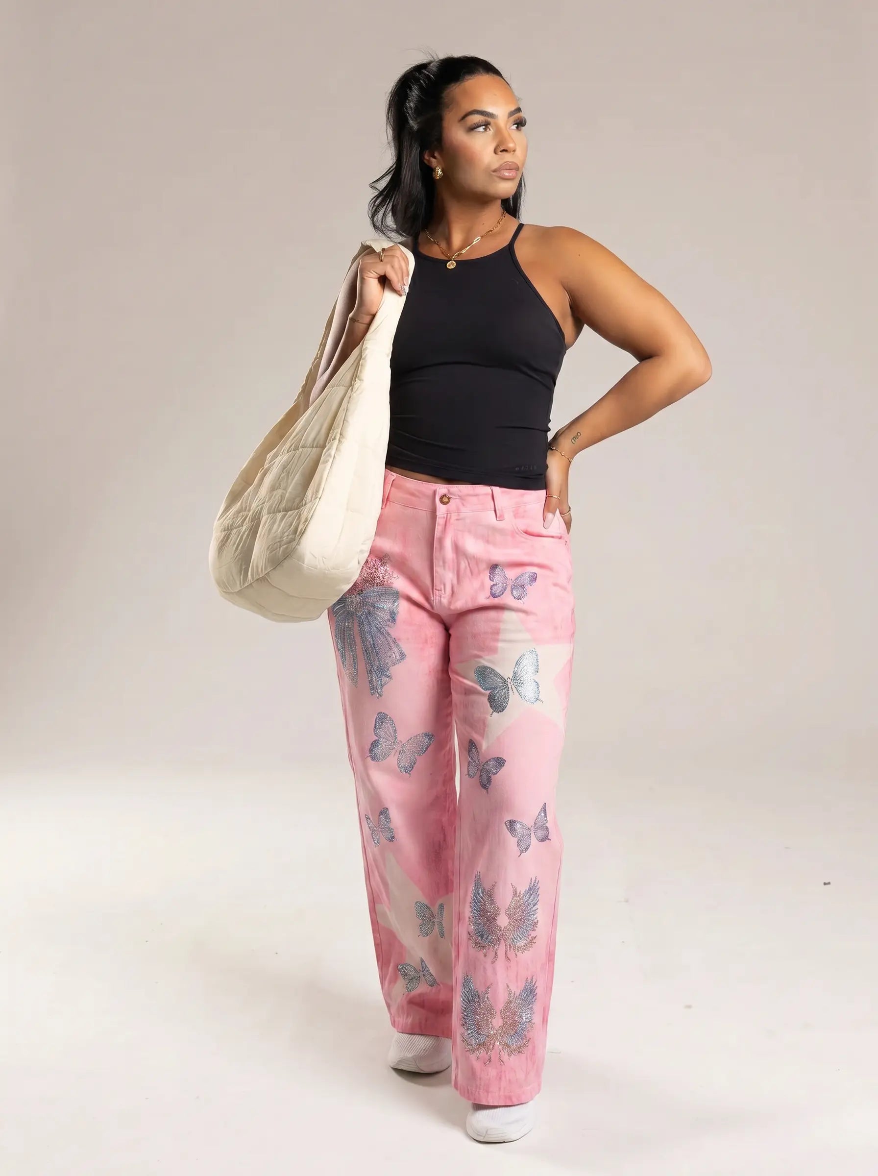 A woman modeling pink wide-leg jeans with a blue butterfly print, paired with a black halter top and carrying a large cream canvas tote bag against a plain studio background. TCH Pink Butterfly Rhinestone Star Wide Leg Jeans