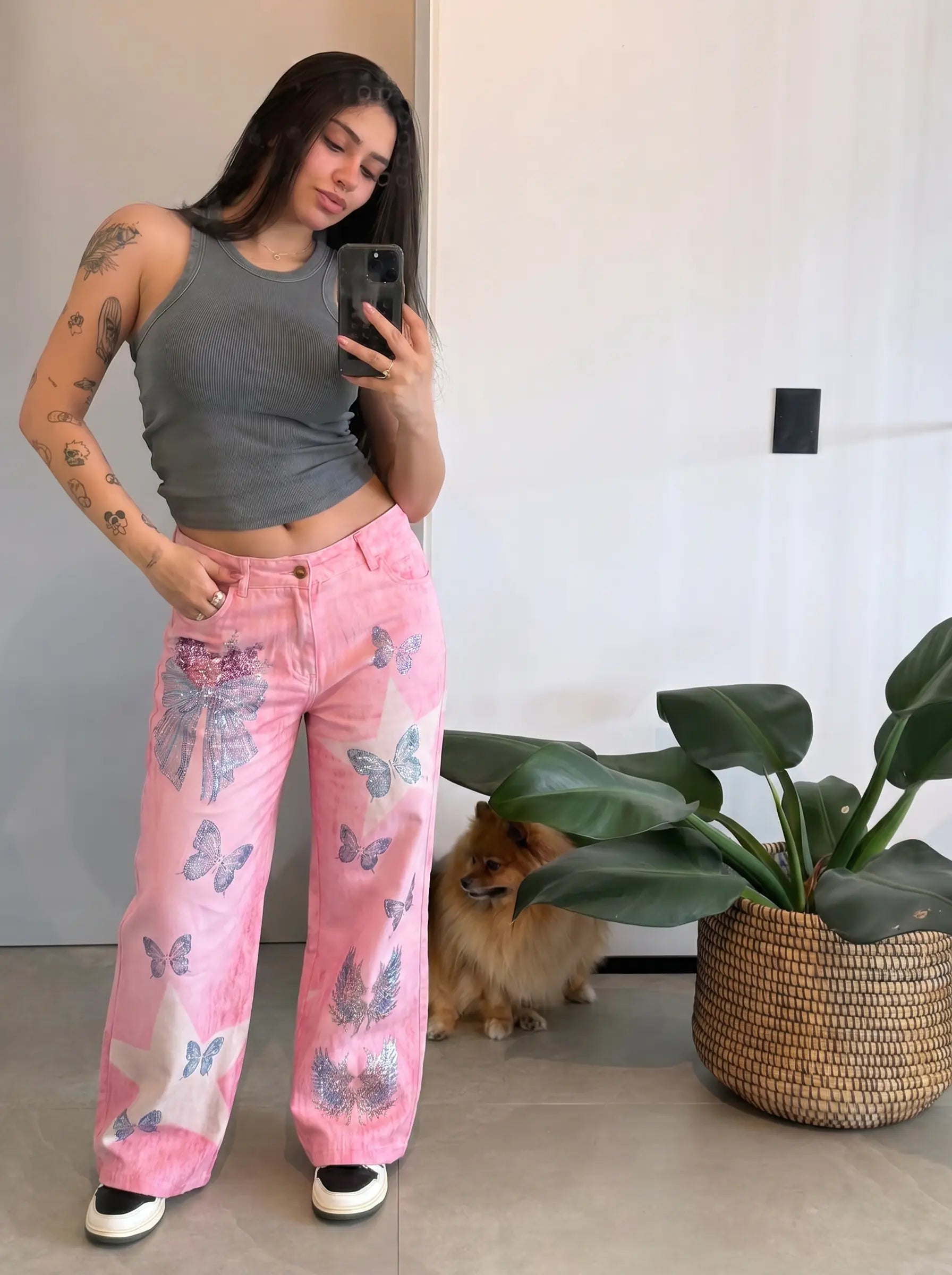 A woman takes a mirror selfie wearing pink wide-leg jeans with a butterfly print, a gray ribbed tank top, and black-and-white sneakers. A small dog sits on the floor next to a potted green plant in a woven basket, against a plain indoor wall. TCH Pink Butterfly Rhinestone Star Wide Leg Jeans