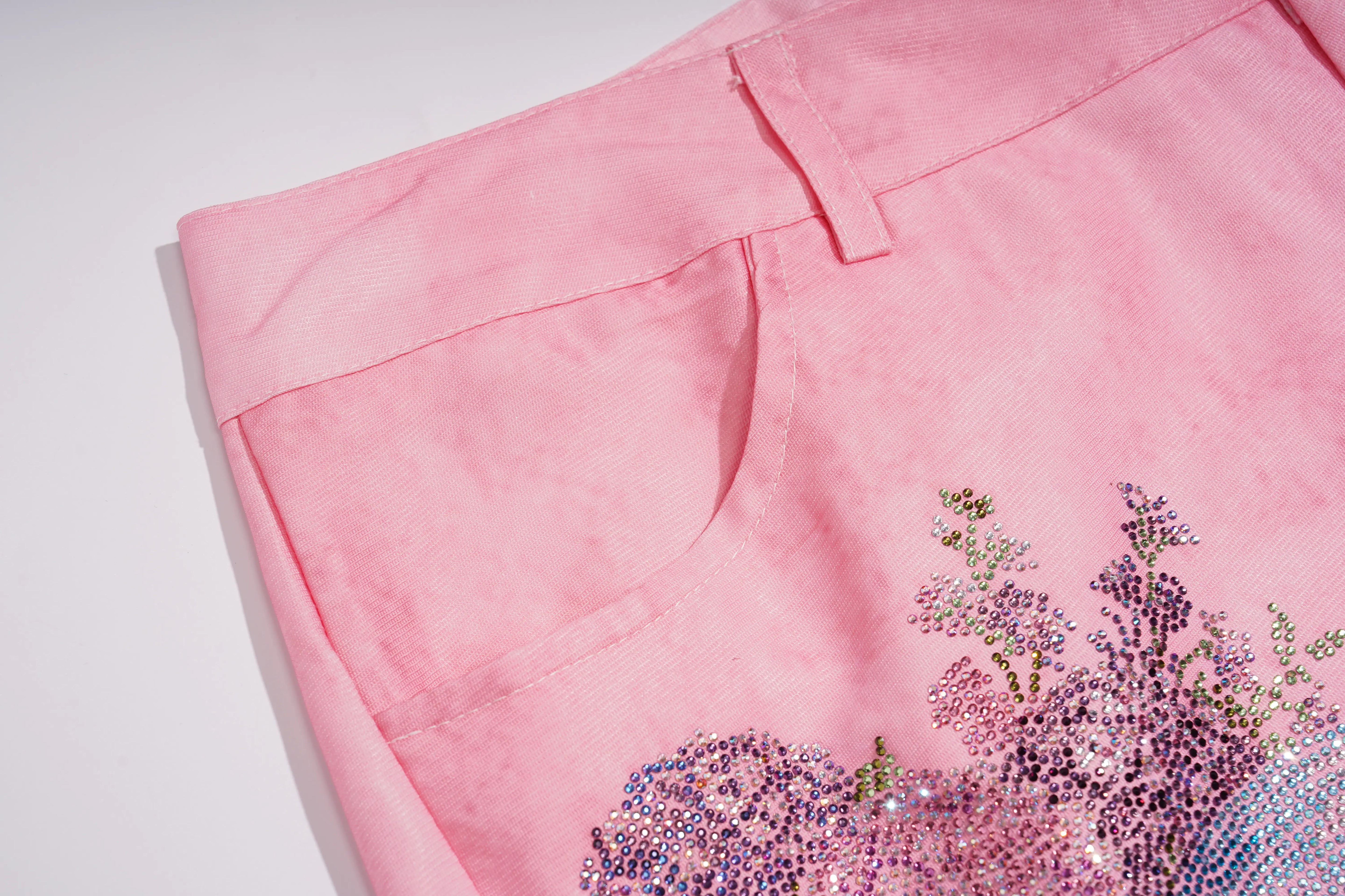 A close-up of a pink skirt or shorts showing the waistband, belt loop, and a decorative floral sequin embellishment in purple, blue, and pink near the lower right hem, set against a plain white background. TCH Pink Butterfly Rhinestone Star Wide Leg Jeans