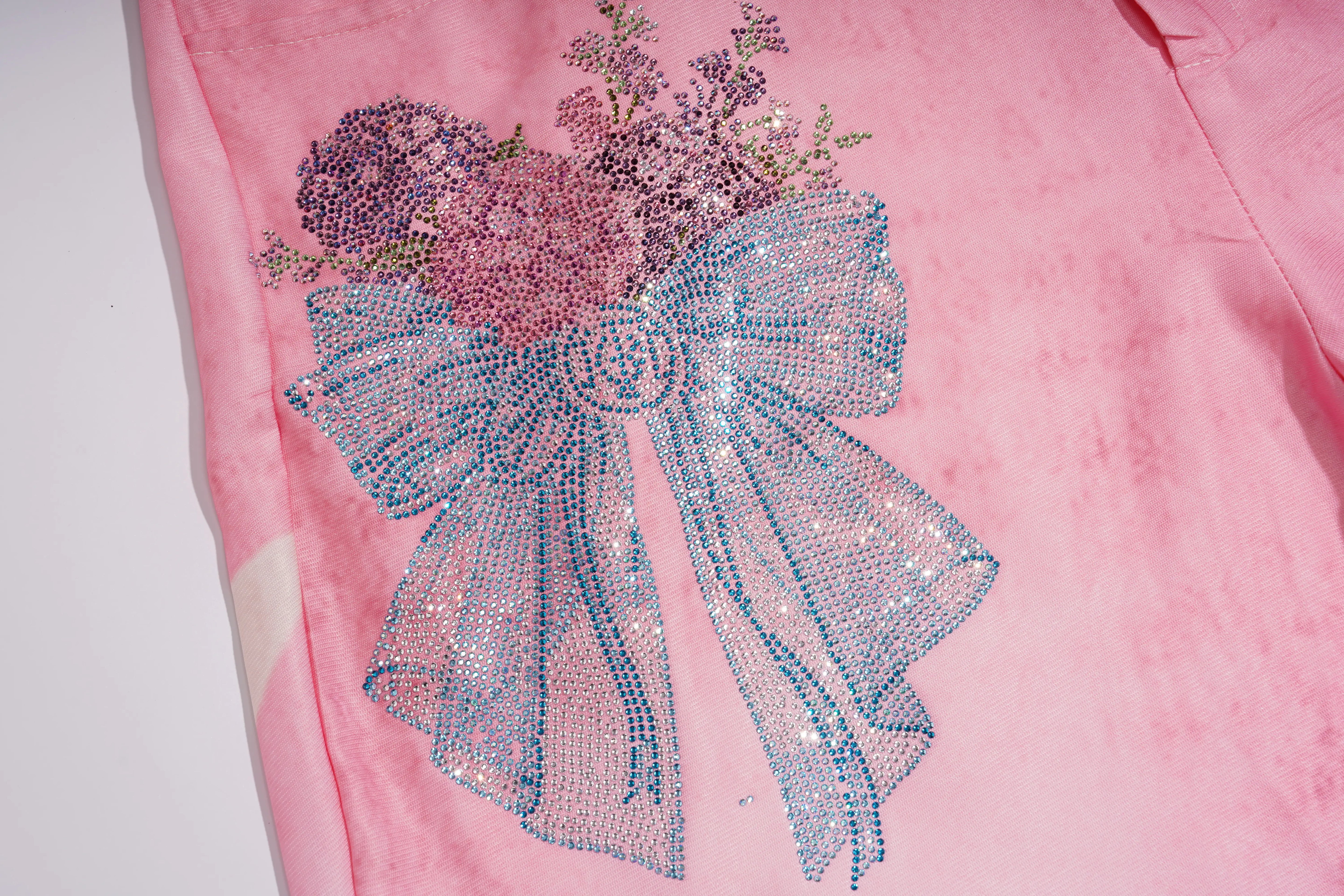 A close-up of a pink garment featuring a large rhinestone bow and floral bouquet applique in shades of blue, purple, and pink. TCH Pink Butterfly Rhinestone Star Wide Leg Jeans