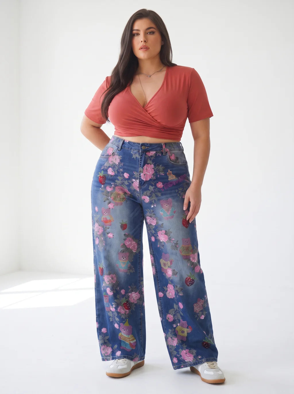 A woman modeling a coral wrap-style short-sleeve crop top and high-waisted wide-leg blue jeans with a colorful floral and whimsical print, paired with white sneakers, in a bright white studio setting. TCH Pink Floral Teddy Bear Graphic Wide-Leg Jeans