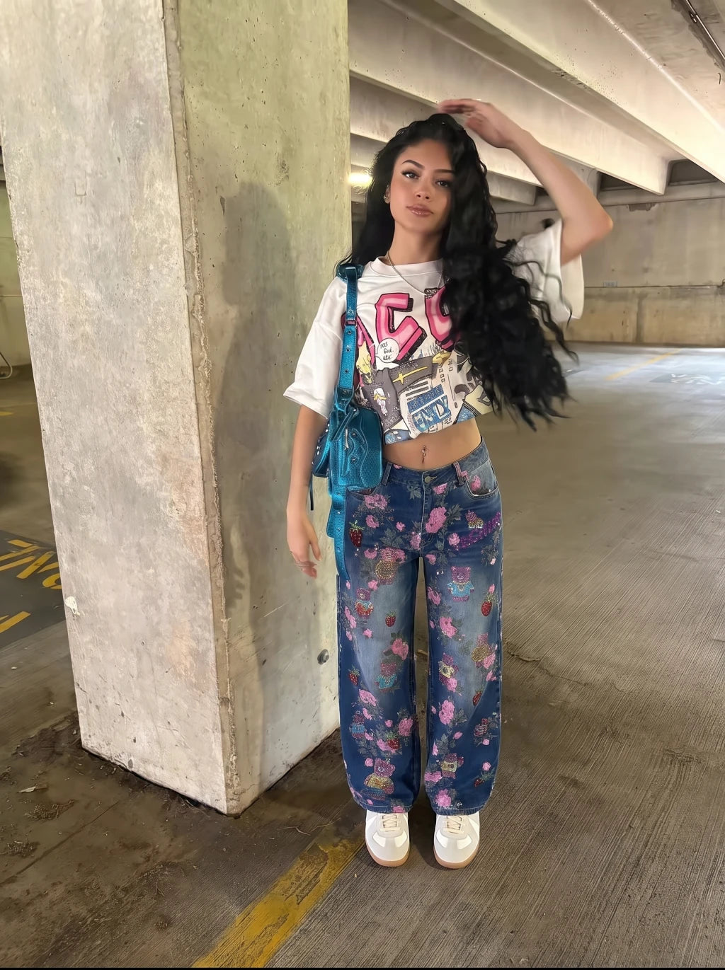 A woman stands in a concrete parking garage wearing a white graphic crop top, blue floral wide-leg jeans, white sneakers, and a blue shoulder bag. TCH Pink Floral Teddy Bear Graphic Wide-Leg Jeans