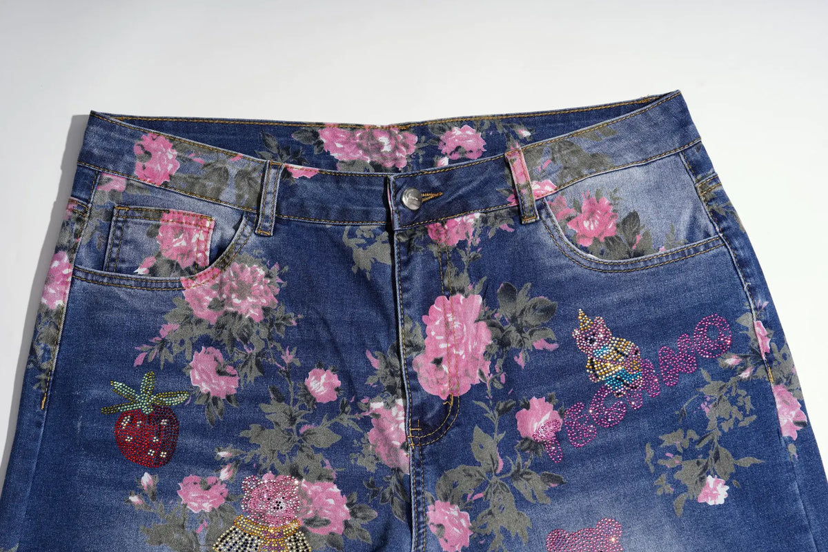 Close-up of blue denim jeans with a pink floral print, embellished with a strawberry patch, a butterfly patch, and rhinestone lettering on the right leg, shown against a plain white background. TCH Pink Floral Teddy Bear Graphic Wide-Leg Jeans