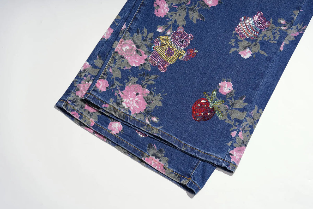 A folded pair of blue denim jeans featuring a pink floral print and embellished with colorful rhinestone patches, including a teddy bear and a strawberry, shown on a plain white background. TCH Pink Floral Teddy Bear Graphic Wide-Leg Jeans
