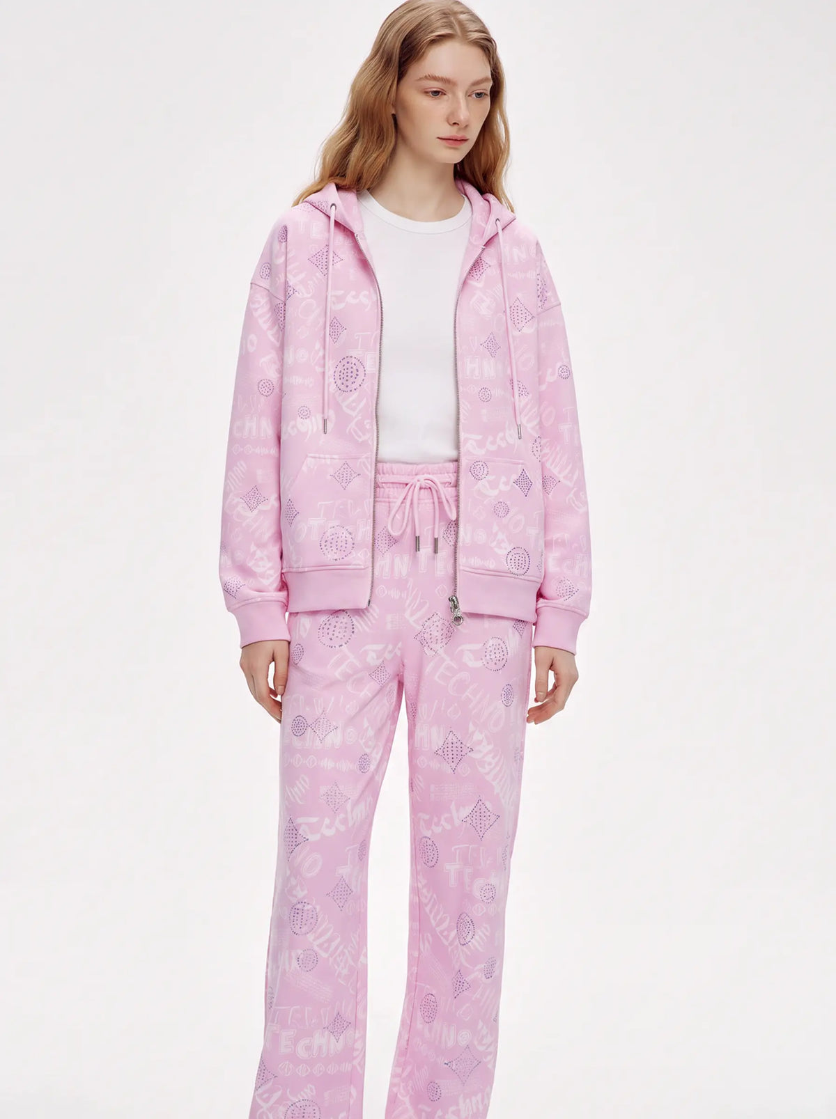 A model wearing a light pink two-piece tracksuit with a subtle tonal graphic pattern, consisting of a zip-up hoodie and matching sweatpants, styled over a white t-shirt against a plain white studio background. TCH Pink Monogram Graphic Zip Up Hoodie