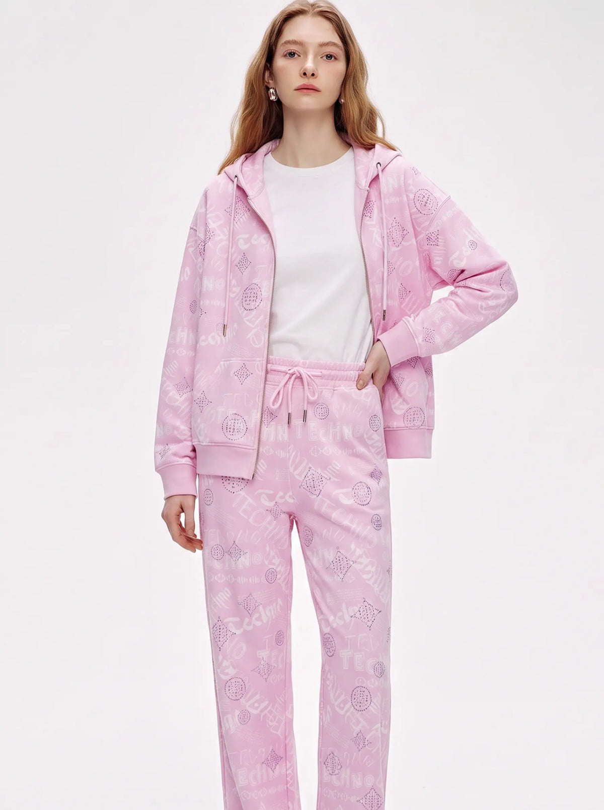 A woman modeling a light pink two-piece tracksuit with a subtle tonal graphic pattern, consisting of a zip-up hoodie and matching sweatpants, worn over a white t-shirt against a plain white studio background. TCH Pink Monogram Graphic Zip Up Hoodie