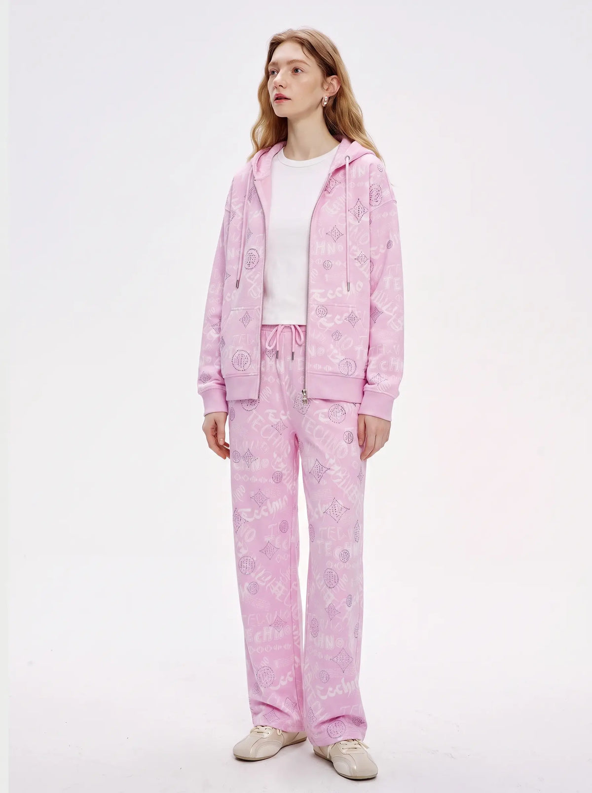 A woman modeling a light pink two-piece tracksuit with a subtle tonal monogram pattern, consisting of a zip-up hoodie and matching sweatpants, styled over a white t-shirt and beige sneakers against a plain white studio background. TCH Pink Monogram Graphic Zip Up Hoodie