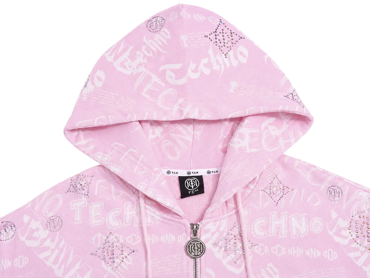 A close-up of a light pink zip-up hoodie featuring an all-over white graffiti-style text and graphic print, with a black and white Tula brand label visible at the inner collar. TCH Pink Monogram Graphic Zip Up Hoodie