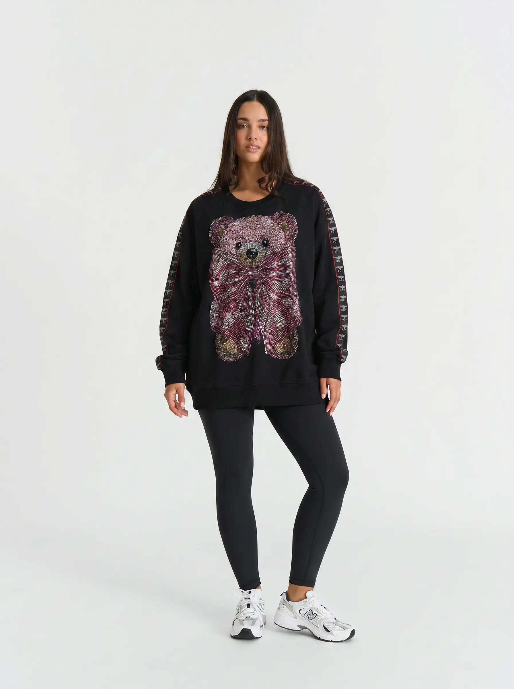A woman modeling a black crewneck sweatshirt featuring a large, colorful sequined teddy bear graphic on the front, paired with black leggings and white sneakers against a plain white studio background. TCH Pink Teddy Bear Rhinestone Graphic Black Sweatshirt