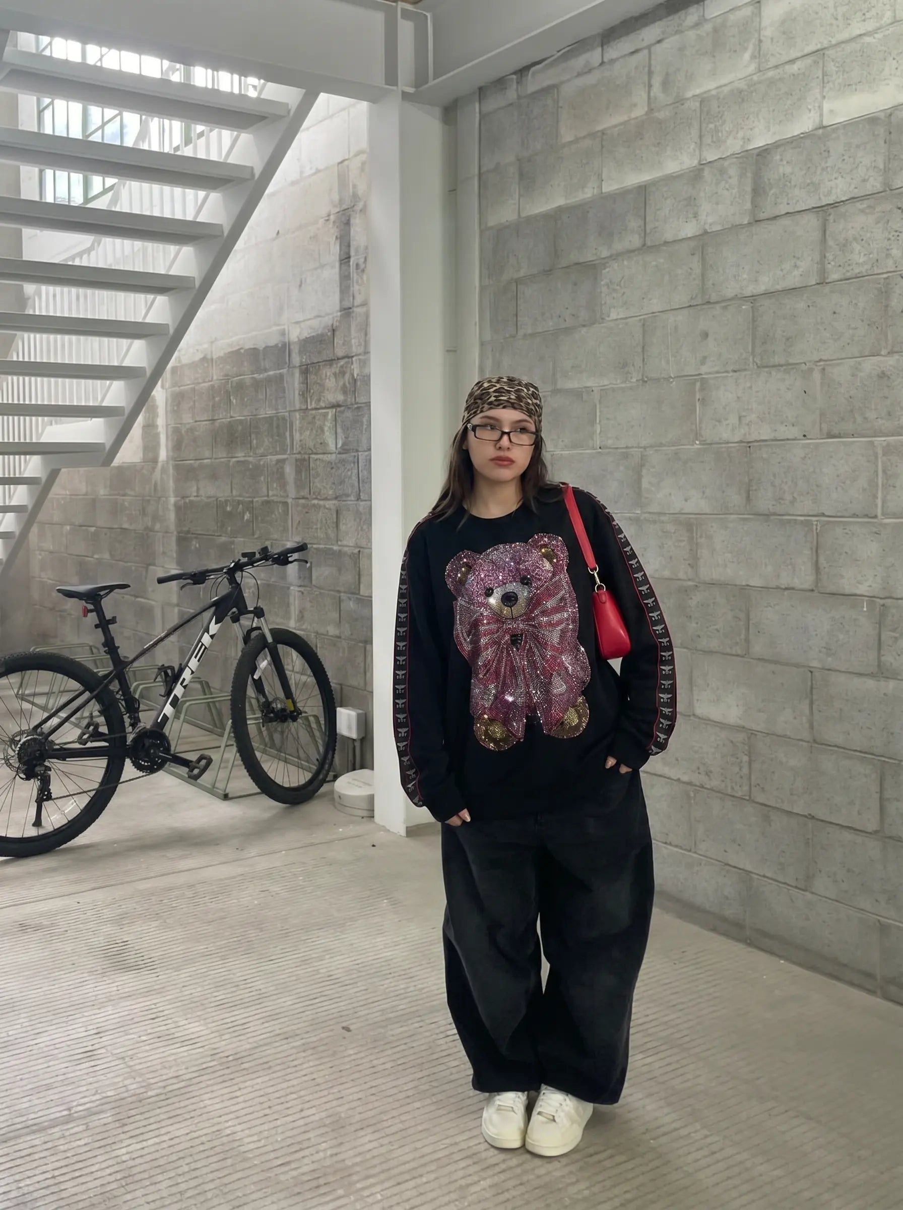 A person wearing a black long-sleeve sweatshirt with a large sequined teddy bear graphic on the front, paired with black wide-leg sweatpants, a patterned beanie, and white sneakers. They are carrying a red shoulder bag and standing in a concrete corridor with a bicycle in the background. TCH Pink Teddy Bear Rhinestone Graphic Black Sweatshirt