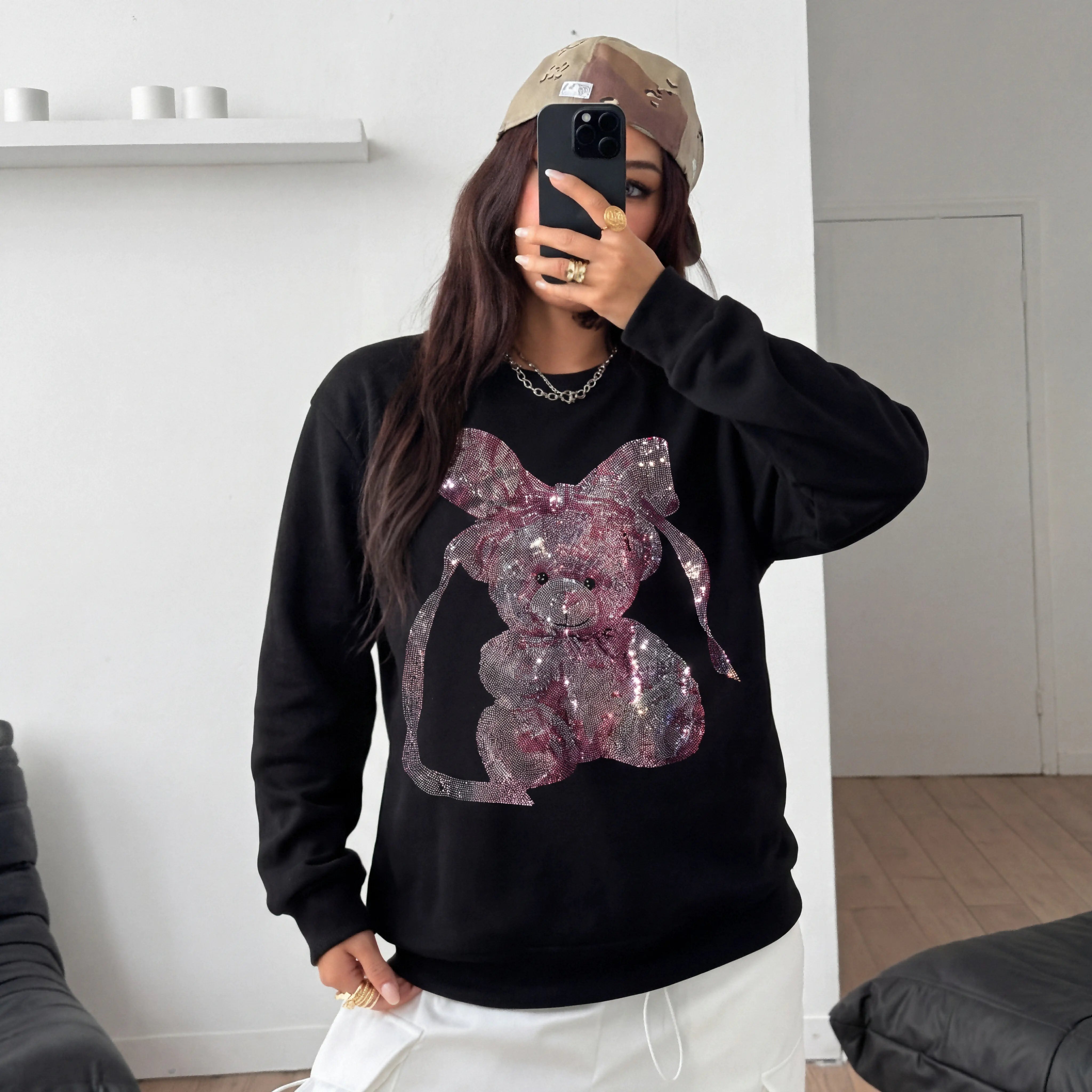 Person wearing a black sweatshirt with a large pink bow design, taking a mirror selfie. TCH Pink Teddy Rhinestone Sweatshirt
