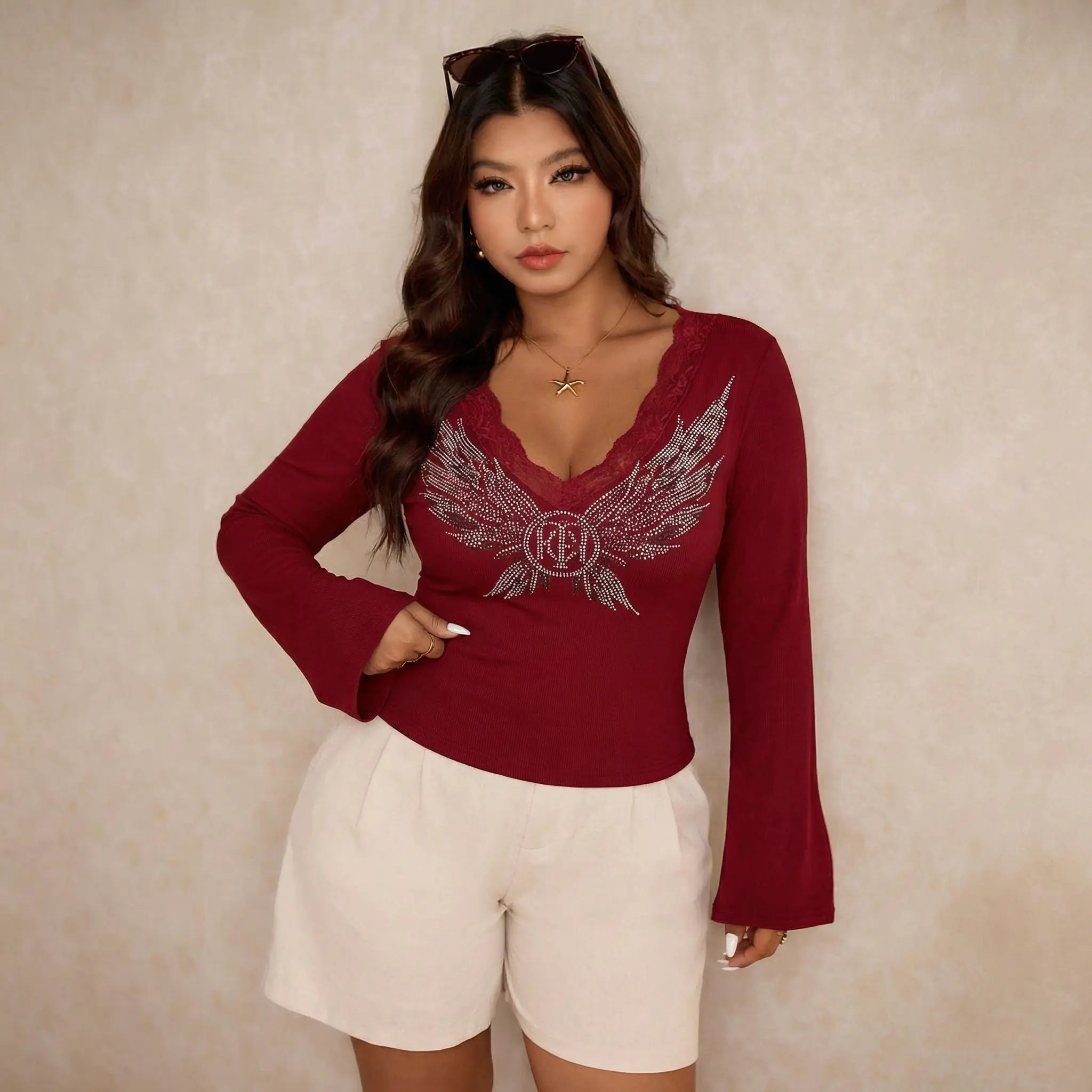 Woman wearing a red long-sleeve top with a logo and white shorts against a beige background, TCH Red Lace Trim V-Neck Rhinestone Wing Long Sleeve Top
